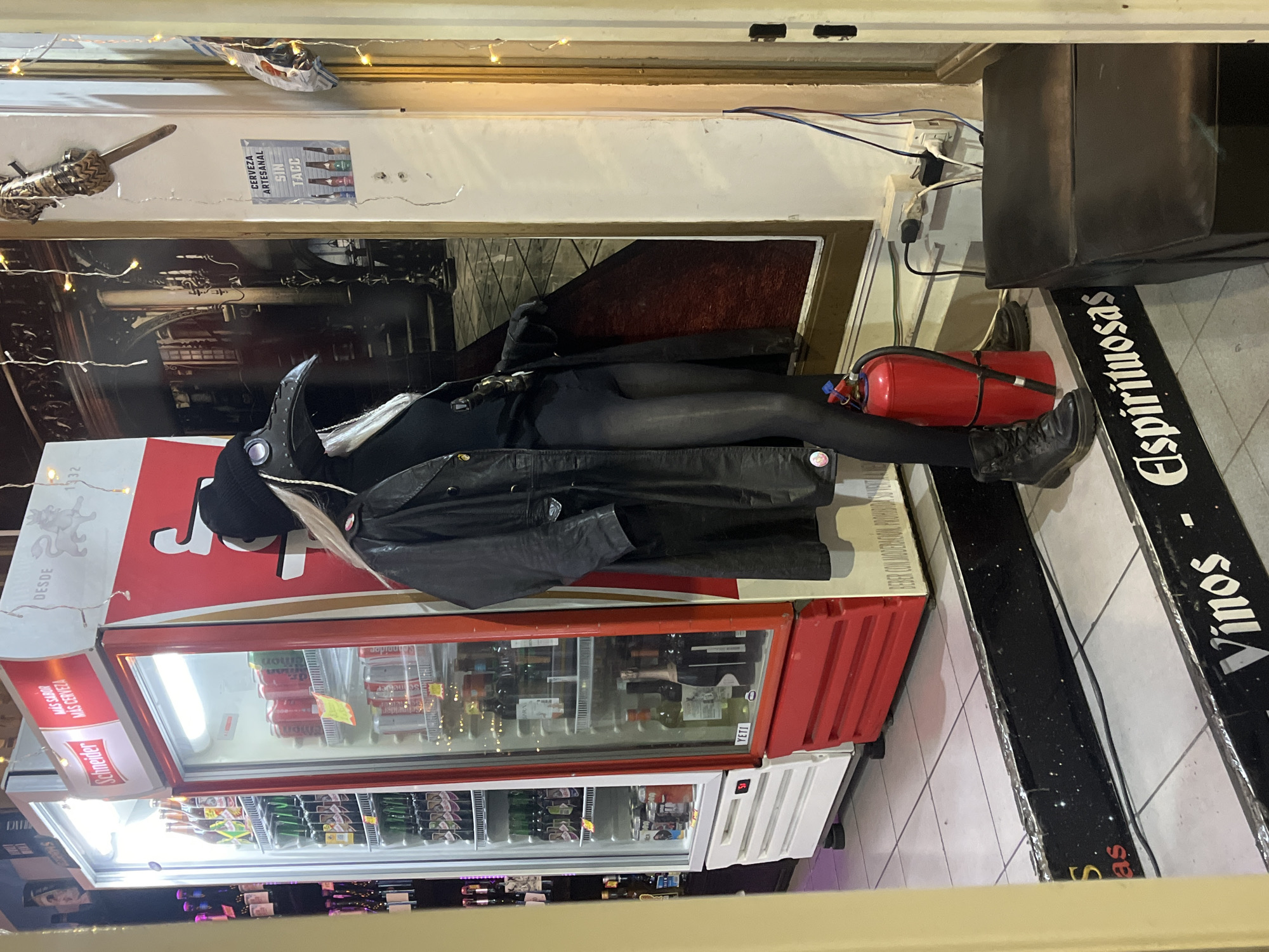 The image shows a person lying on top of a red and white Schneider beer refrigerator in what appears to be a bar or restaurant in Buenos Aires, Argentina. The person is wearing a black outfit with a plague doctor mask and a black leather jacket. Next to the person, there's a red fire extinguisher. A black signboard reading "Vinos - Espirituosas" is adjacent to the refrigerator. The background includes beer advertisements, a depiction of a street, decorative lights, and various objects on shelves.