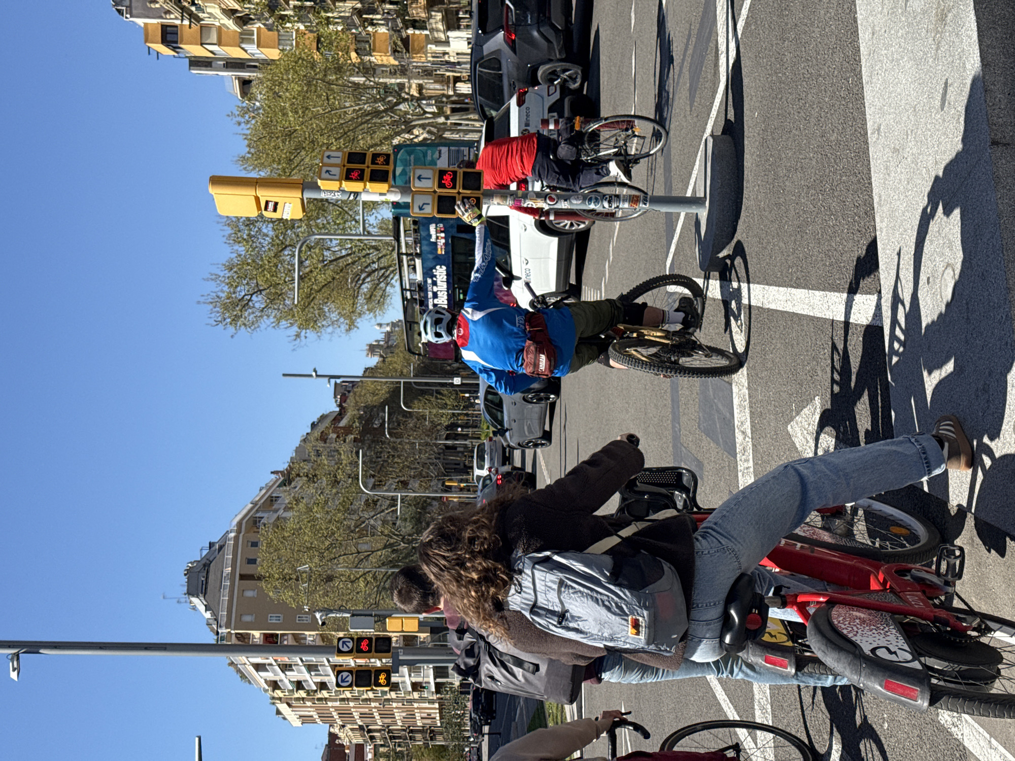 The image captures a street scene in Barcelona, Spain, featuring cyclists and pedestrians at a traffic intersection. The location is suggested by the architectural style of the surrounding buildings, including the typical balconies and design elements common in Barcelona. Several people are on bicycles, waiting at what appears to be a bike-specific traffic signal. One cyclist is signaling with his arm, possibly indicating a turn. Other individuals are walking nearby. Several cars and a double-decker bus are visible in the background, indicating urban traffic. The day is sunny, with shadows clearly visible on the pavement. A traffic light pole with multiple traffic signals and bicycle lanes signs are also prominent.