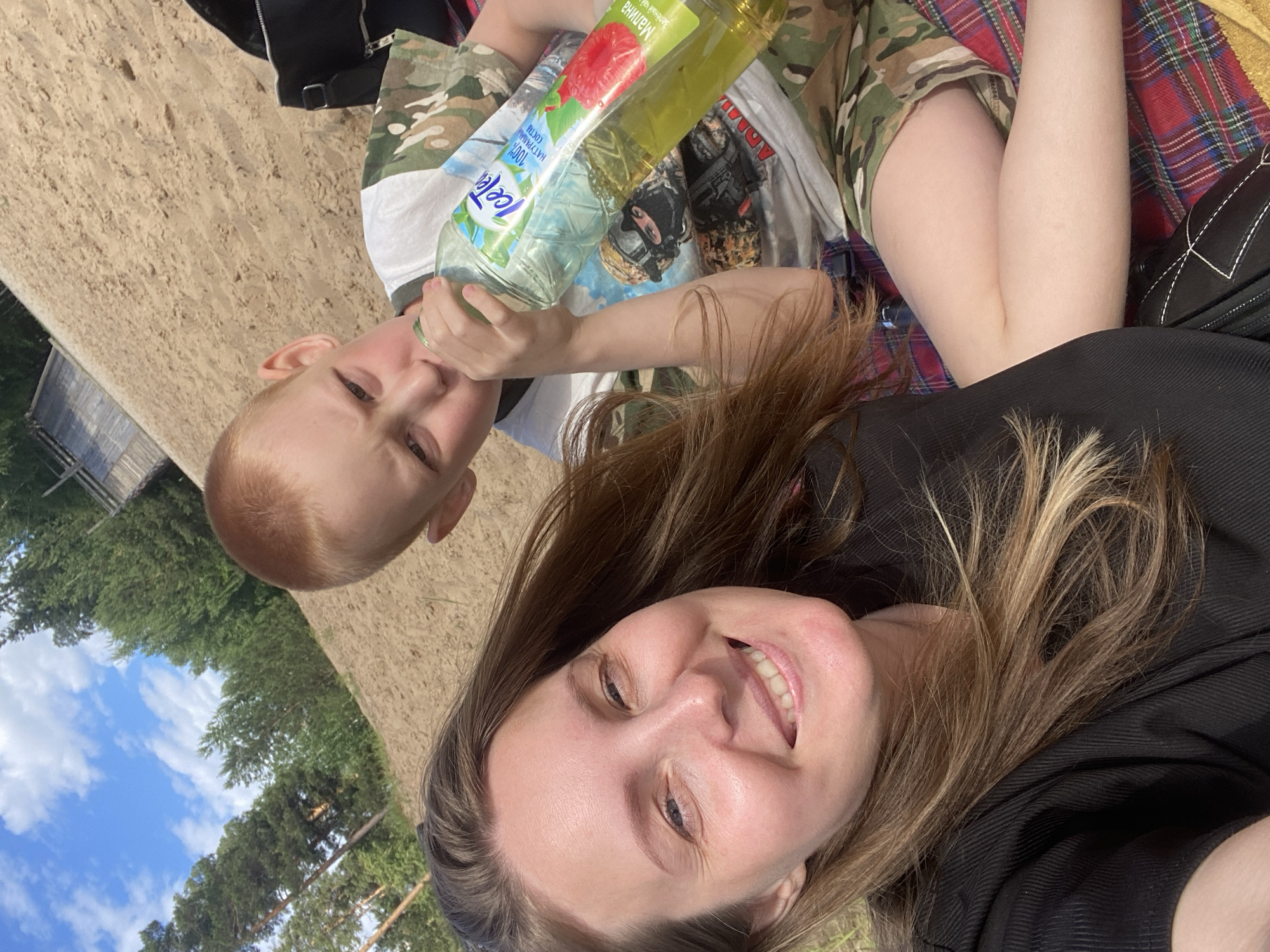 A woman and a young boy are sitting on a red and black checkered blanket on a sandy area. The woman is on the left, smiling at the camera, and has long, light brown hair. She is wearing a black top. The boy is on the right, wearing a camouflage t-shirt and shorts. He is drinking from a large bottle of Ice Tea with a raspberry image on the label. Behind them, there is a sandy slope leading up to trees and a blue sky with white clouds. The location appears to be a park or a recreational area.