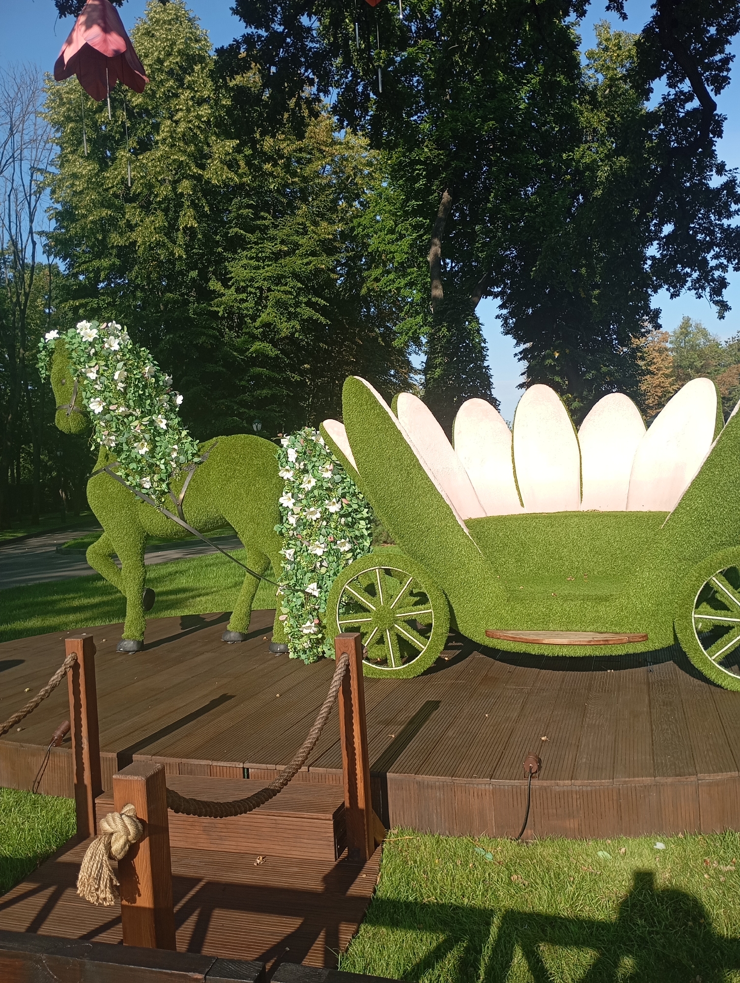 In Kharkiv, Ukraine, a decorative display features a green, topiary horse, intricately adorned with white flowers, harnessed to a flower-shaped carriage. Both are positioned on a wooden platform set amidst lush green grass and mature trees. The display appears to be outdoors, possibly in a park or public garden, bathed in sunlight.