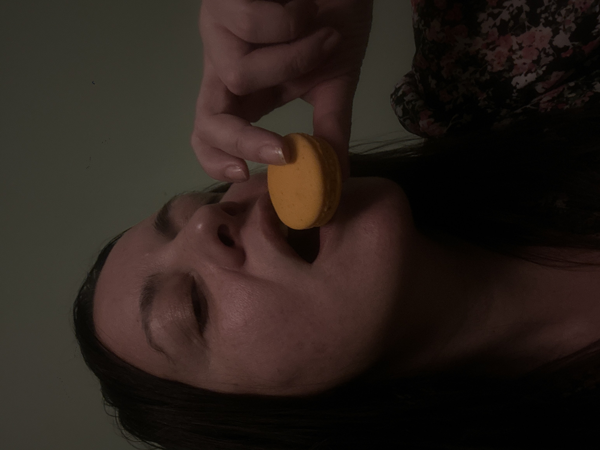 A pale-skinned woman with dark hair is lying on her back. Her eyes are closed, and an orange macaron is being held between someone's fingers and placed in her mouth. The person holding the macaron is wearing a floral-patterned shirt. The background appears to be a muted green. No evidence is present in the image to suggest this image was taken in Riga, Latvia.