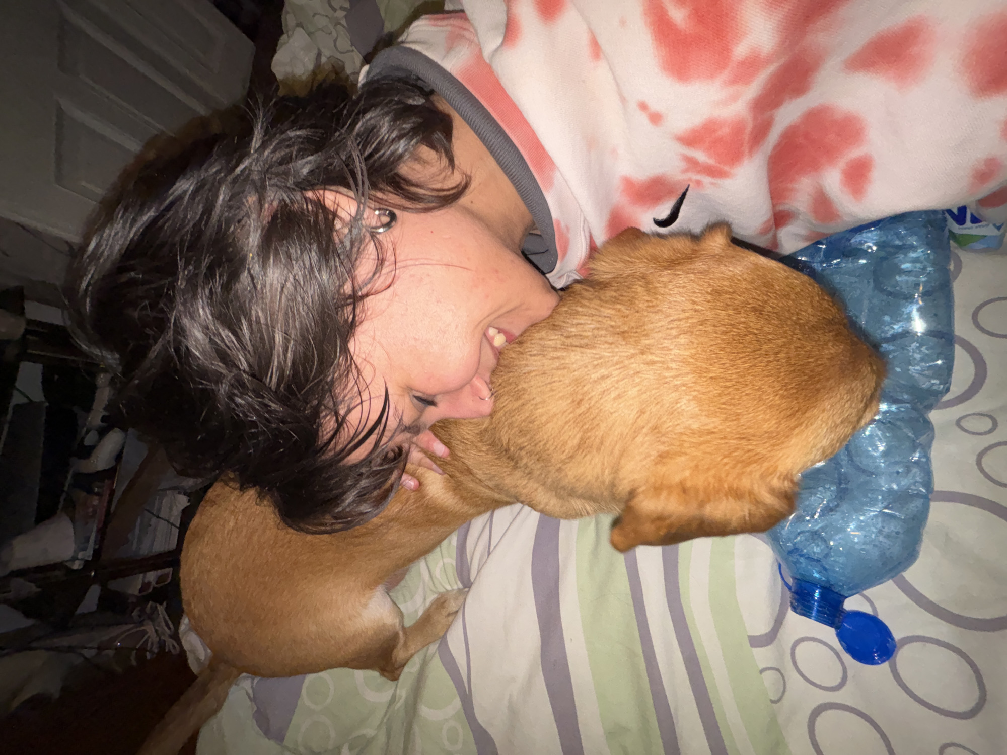 A person with dark, messy hair is lying on a bed, nuzzling a tan-colored dog. The person is wearing a white hoodie with pink tie-dye patterns and a grey collar. They have an earring in their left ear. The dog is positioned on top of a blue plastic bottle filled with a clear liquid and small bubbles. The bed has striped sheets in green, grey, and white. The background is dimly lit and appears to be a bedroom with clothes piled in a corner and a white door. The image was taken indoors.