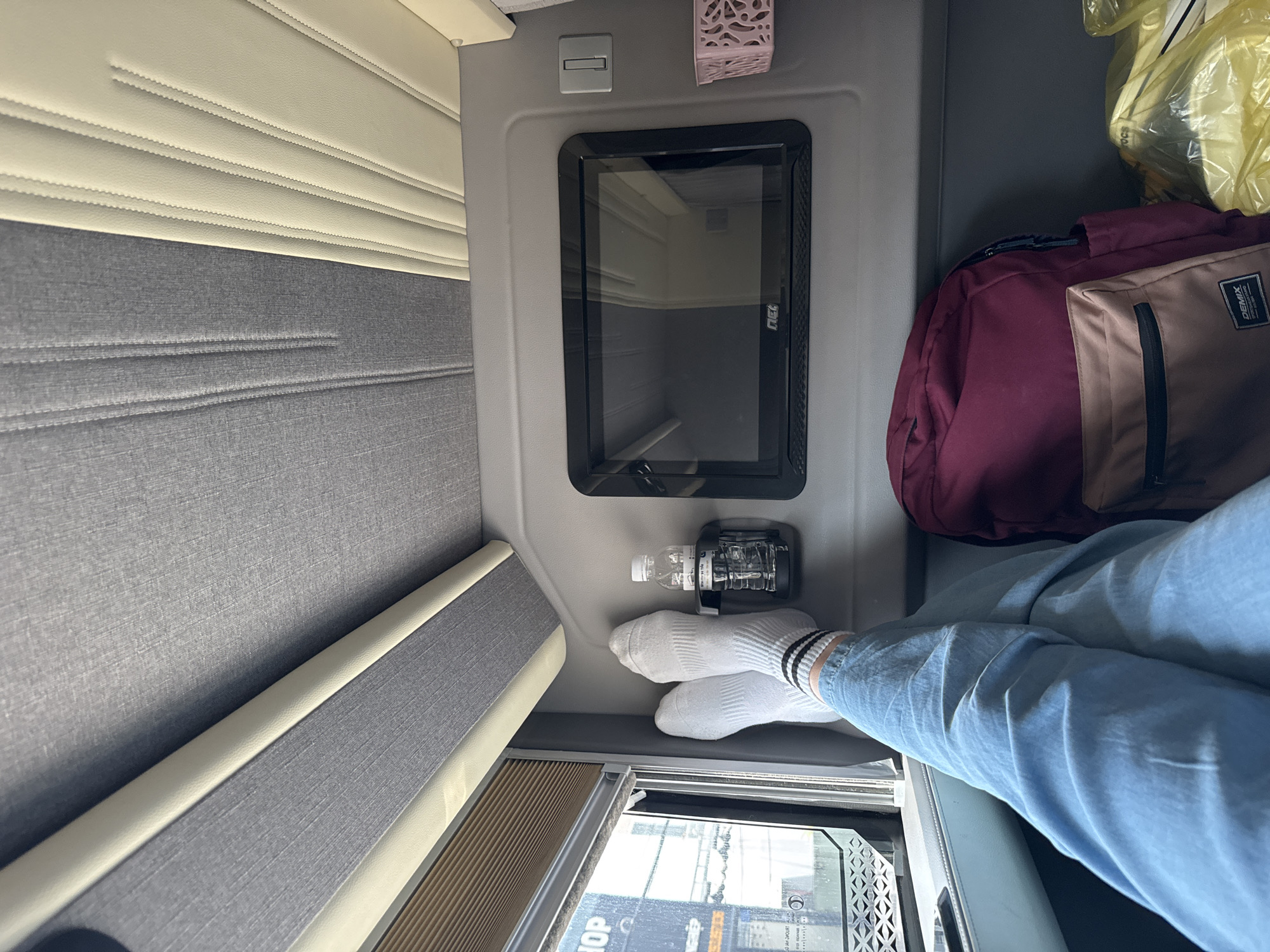 The image captures a person's legs and feet, clad in white socks with black stripes and light blue jeans, resting in what appears to be a seat on a mode of transportation, likely a bus or train, given the interior design. The setting is oriented towards the viewer's left, with a window visible at the bottom of the frame showing a slightly blurred outdoor scene, hinting at daylight.
The interior features a combination of textured grey and beige upholstered surfaces, characteristic of passenger seating areas. Above, a small black framed screen is integrated into the paneling, and a decorative pink item, possibly an air freshener, is attached to the wall. A small bottle of water is secured in a holder next to the screen.
In the background, a dark red backpack and a yellow plastic bag are visible, suggesting personal belongings. The overall mood is relaxed and casual, with the focus on the resting posture of the individual. The visible text on the backpack reads "DEMIX". The location context provided is Thủ Đức, Vietnam.