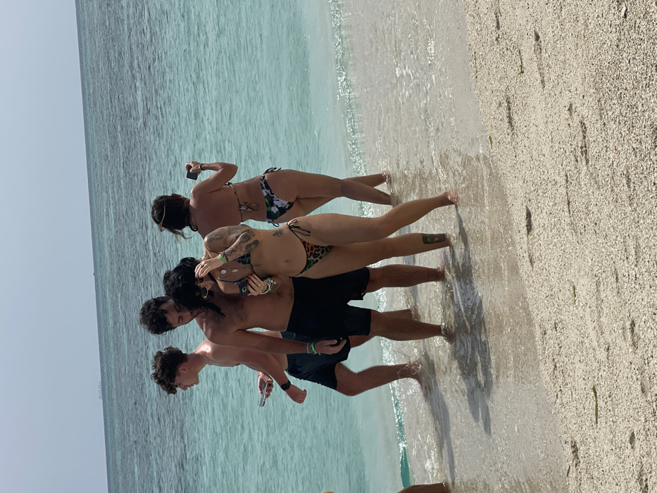 The image displays four individuals standing in the shallow, clear turquoise-blue ocean water at a beach in Zanzibar, Tanzania. The foreground shows light-colored sand, partially wet from the incoming waves.
From left to right:
1. A male, bare-chested and wearing dark swim shorts, stands with his left side partially facing the camera, looking towards the center of the group. He holds a silver smartphone in his left hand.
2. A second male, bare-chested and wearing dark swim shorts, stands facing slightly left, looking down towards the female in the leopard print bikini. He has dark hair and a green wristband on his right arm.
3. A female, wearing a leopard print string bikini, stands with her body slightly angled towards the camera, looking down at a smartphone held in both hands. She has numerous tattoos on both arms, including a visible sleeve on her left arm, and a tattoo on her right calf. She wears multiple necklaces and a light-colored wristband on her right wrist.
4. A second female, with her back mostly to the camera, stands looking out at the ocean. She wears a dark bikini with a white and green floral pattern and holds a dark smartphone horizontally, appearing to be taking a photo or video of the sea.
The water level reaches the individuals' ankles and lower calves. In the distant background, a faint structure is visible on the horizon of the ocean under a pale blue sky.