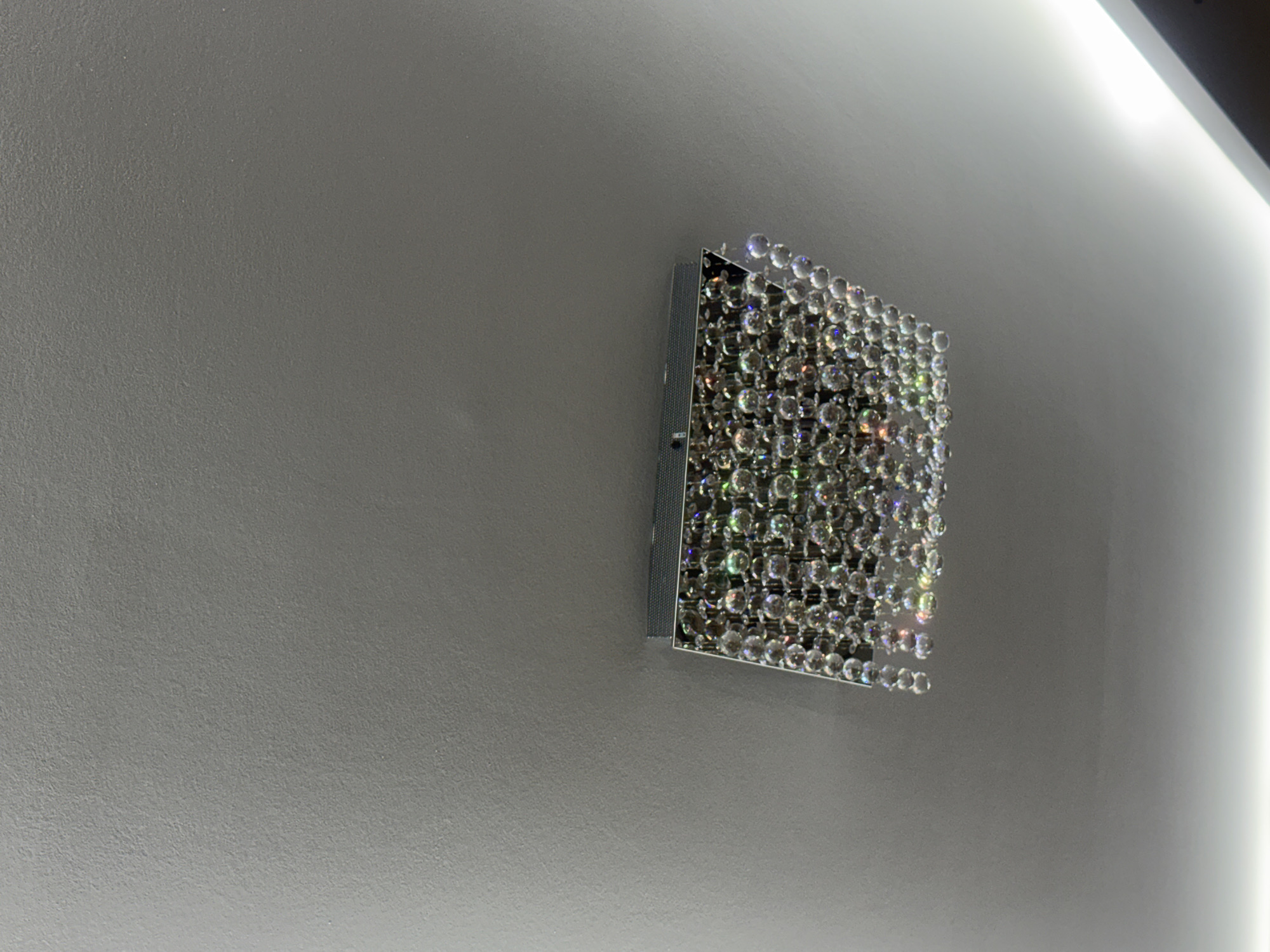 A square, possibly reflective, object adorned with small, round, clear gems or crystals is affixed to a gray wall. The gems capture and reflect light, creating a sparkling effect. The object is positioned off-center, towards the right side of the frame. The gray wall has a smooth texture. A bright, linear light source is visible along the right edge of the image, indicating either a lighting fixture or sunlight streaming in. The environment is indoors. The location is unknown as it cannot be determined from the image.