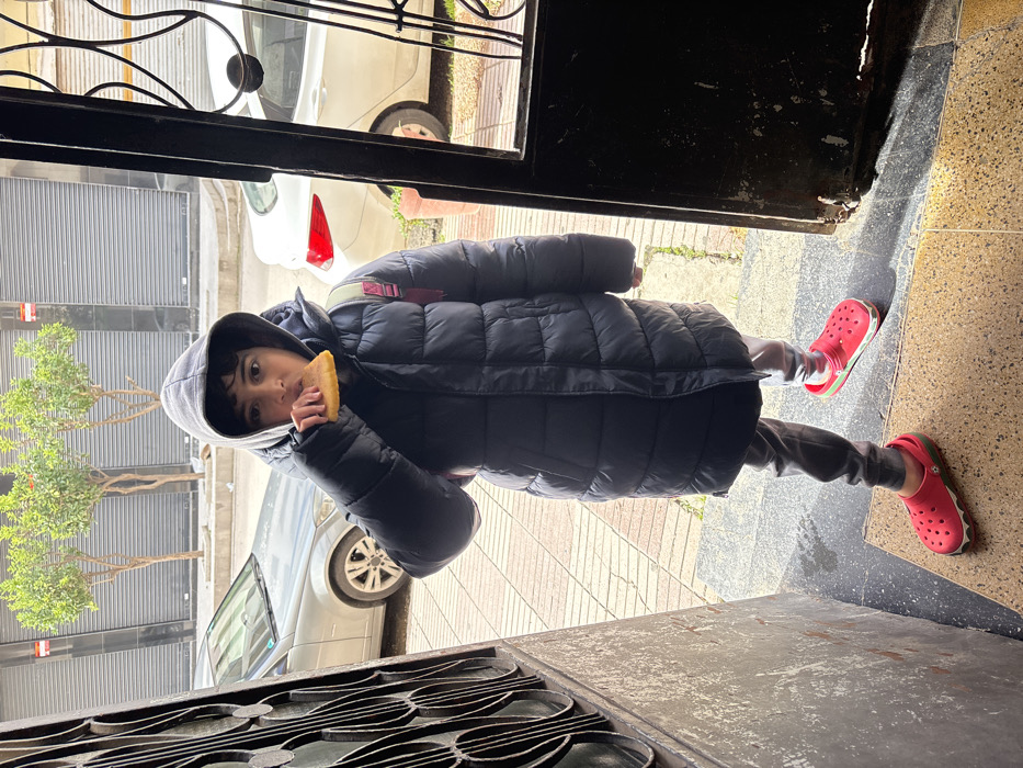 A child stands centered at an open doorway in Meknes, Morocco. The subject, wearing a dark hooded puffer coat, dark trousers, and red Crocs-style clogs with green soles, holds a piece of yellowish, flat food to their mouth with their right hand, appearing to be mid-bite. A pink backpack strap is visible over the child's right shoulder. The child's gaze is directed forward, towards the viewer.
The doorway opens to an outdoor urban environment. Immediately outside, a paved sidewalk features light-colored pavers. Beyond the sidewalk is a street where two cars are parked: a silver Mercedes-Benz sedan is partially visible on the left, and a white sedan's rear is visible on the right. Further in the background, three small trees with green foliage stand in planters along the sidewalk. Behind the trees and cars, building facades are visible, characterized by dark, corrugated metal roll-down security shutters. The interior foreground features light-colored, speckled floor tiles. To the immediate left of the child, a dark, ornate metal grille or gate is partially visible. The lighting is bright and diffuse, indicative of an overcast sky.