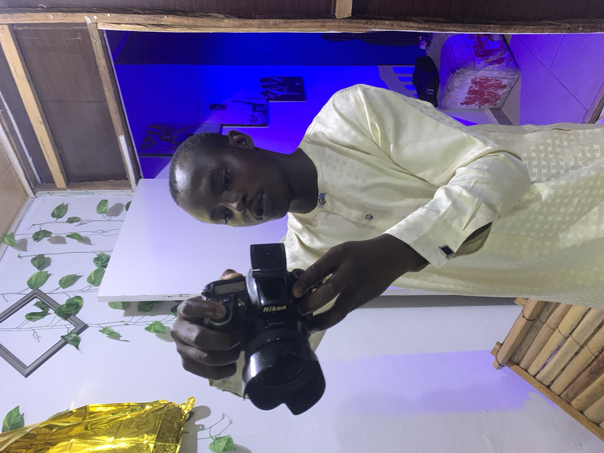 A man stands inside a room and adjusts a Nikon camera. The walls are decorated with fake vines, a picture frame, and some gold wrapping. A white shelf sits behind him, and a blue light illuminates the upper portion of the background. An unfinished ceiling is visible. The image does not provide enough information to determine that it was taken in Jos, Nigeria.