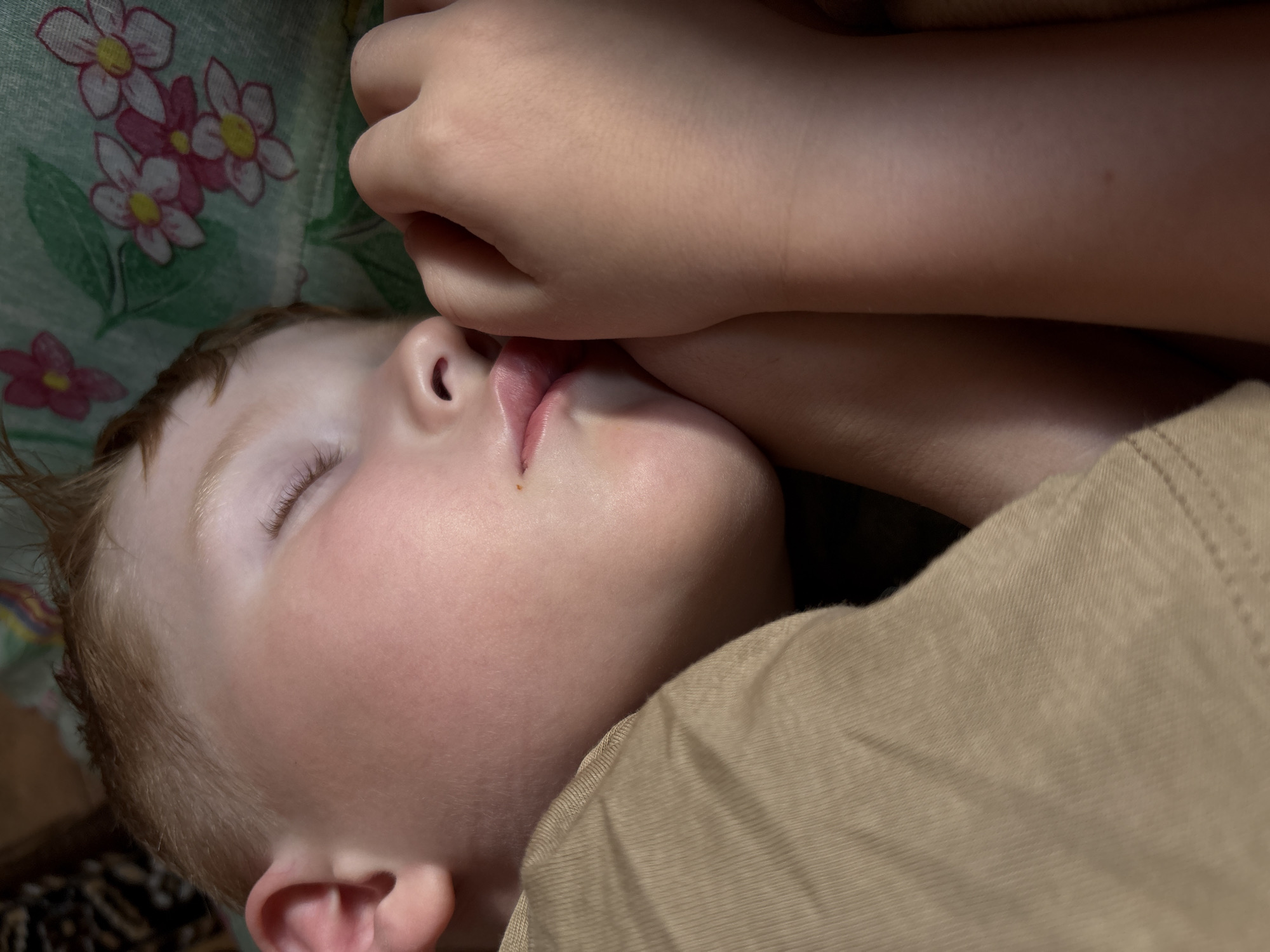 The image shows a close-up, overhead view of a young child sleeping. The child's eyes are closed, and their head is turned to the side. A hand is resting near the child's face, with fingers gently touching their cheek. The child is wearing a light-colored t-shirt. The background features a fabric with a floral pattern in shades of green, pink, and yellow, possibly a blanket or pillow. The lighting suggests it is daytime, possibly indoors. The image conveys a sense of peace and rest. There is no visible text. The location context provided is Italmas, Russia, but there are no specific visual cues in the image to confirm this location.
