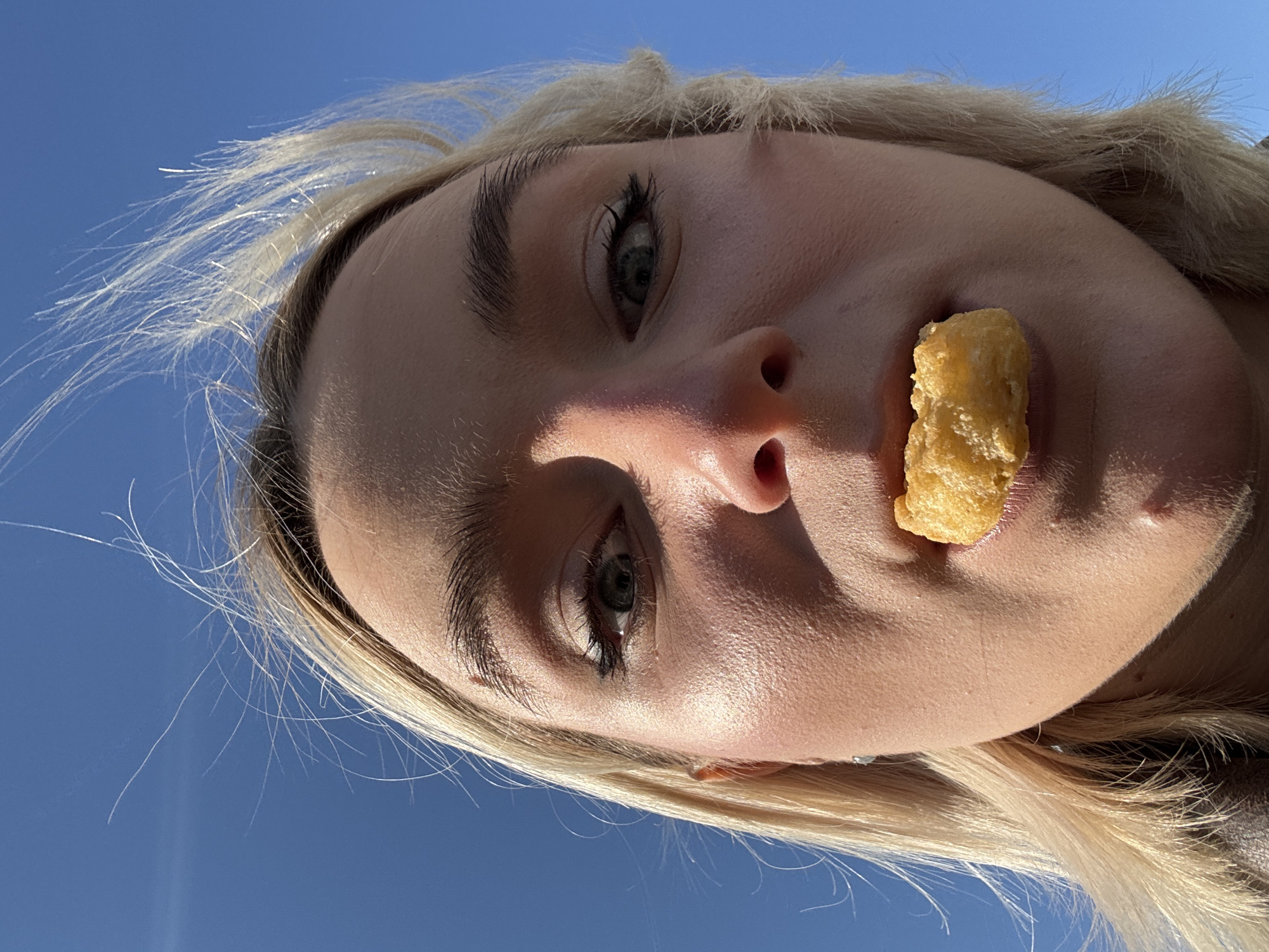 A low-angle close-up shot shows a young woman with blonde hair holding a fried food item against her upper lip. The background is a clear blue sky. The woman has fair skin, visible eyelashes, and dark eyebrows. The food item is rectangular and appears to be deep-fried. There are no buildings or landmarks visible to identify the location as Vsevolozhsk, Russia.