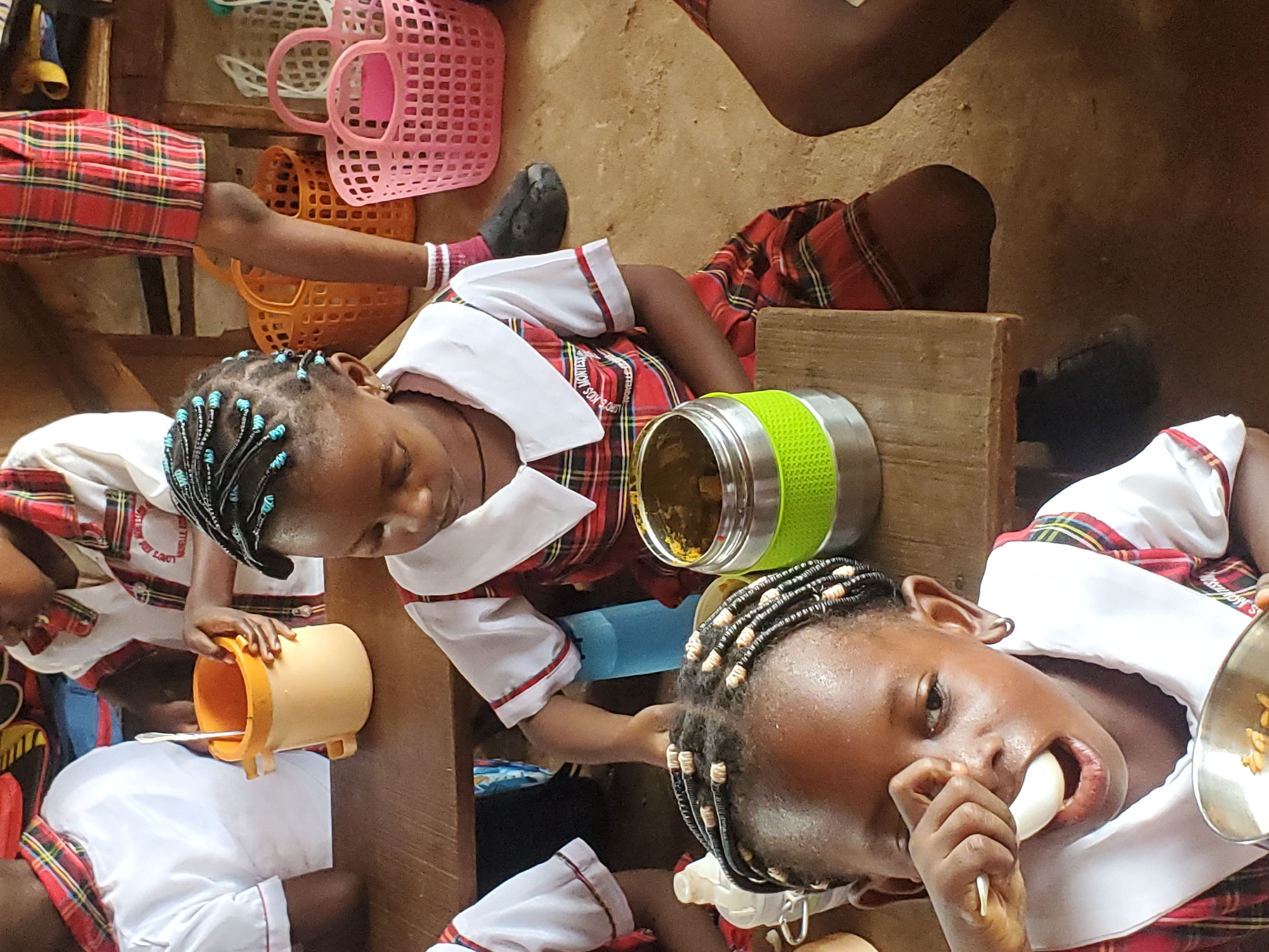 Captured from an elevated perspective, the scene unfolds at an unconfirmed location in Nsugbe, Nigeria. Four children, dressed in matching school uniforms, are eating. The uniforms are composed of red and white plaid dresses with white collars and sleeves trimmed with red. The children have dark skin and intricately braided hair, some adorned with light-colored beads. One child is actively eating from a metal bowl, while another examines the contents of a yellow bucket. A stainless steel lunch container, wrapped with a green material, sits atop a wooden surface, near another child. Pink and orange plastic baskets are stored above, providing additional storage. The background is simple, consisting of dirt and a wall, highlighting the basic environment.