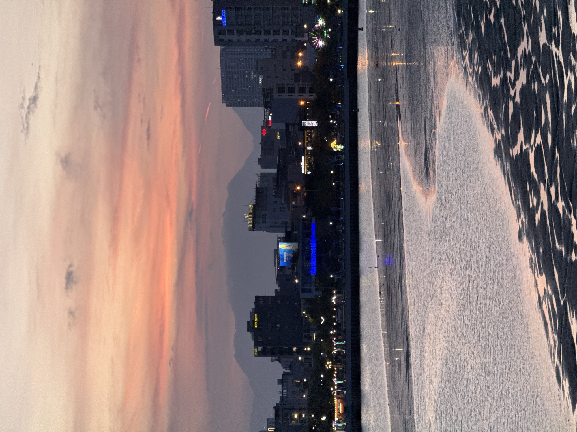 This is an image of the Han River in Da Nang, Vietnam, at sunset. The sky is painted with hues of pink, orange, and purple, with scattered clouds. In the background, the city skyline is visible, featuring tall buildings and mountains in the distance. The riverfront is illuminated by streetlights and colorful decorations, and a ferris wheel can be seen on the right side of the frame. The foreground is dominated by the water, with reflections of the city lights and the sky shimmering on its surface. The exposed riverbed, with its intricate patterns of sand and mud, adds texture and depth to the scene.