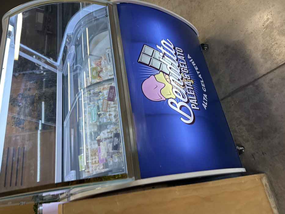 A refrigerated display case filled with frozen treats on sticks is shown. The case is curved with glass panels. It's decorated with a blue background and a logo featuring a chocolate bar and an ice cream cone. The logo says "Bendito, Paleta y Gelato, Alta Gelateria." The case rests on a wooden counter. A gray floor is visible in the background.