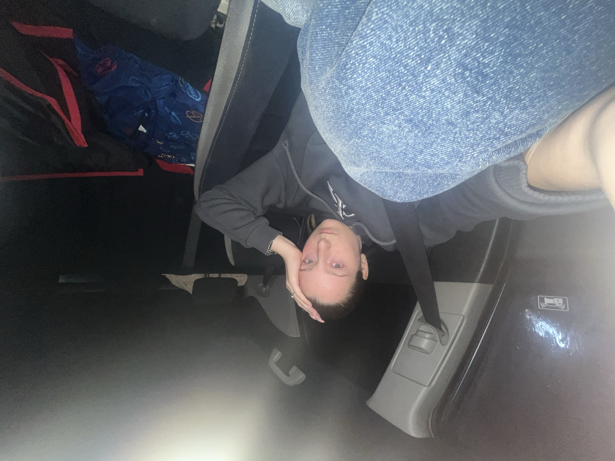 The image shows a young person, likely female, positioned upside down within what appears to be the interior of a vehicle, possibly the back seat or cargo area, in Varna, Bulgaria. She has light skin, dark hair pulled back, and is looking directly at the camera with a neutral to slightly pensive expression. Her right hand (when viewed right-side up) is resting against her forehead.
She is wearing a dark, possibly grey or black, hooded jacket or sweatshirt, and blue denim jeans. On her right hand, she wears a ring, and a bracelet is visible on her left wrist.
The surrounding environment suggests a car interior. To the top-left (when viewed right-side up), a black bag with prominent red straps is visible, along with a bunched-up blue fabric item featuring light-colored patterns. To the right, grey plastic trim and a seatbelt mechanism are clearly seen. A small white pictogram is visible on the grey panel, resembling an ISOFIX or child safety seat anchor symbol. The lighting is dim overall, with a bright light source illuminating the lower-left portion of the frame, creating a gradient from dark to light. The image captures a casual moment or a creative pose within the vehicle.