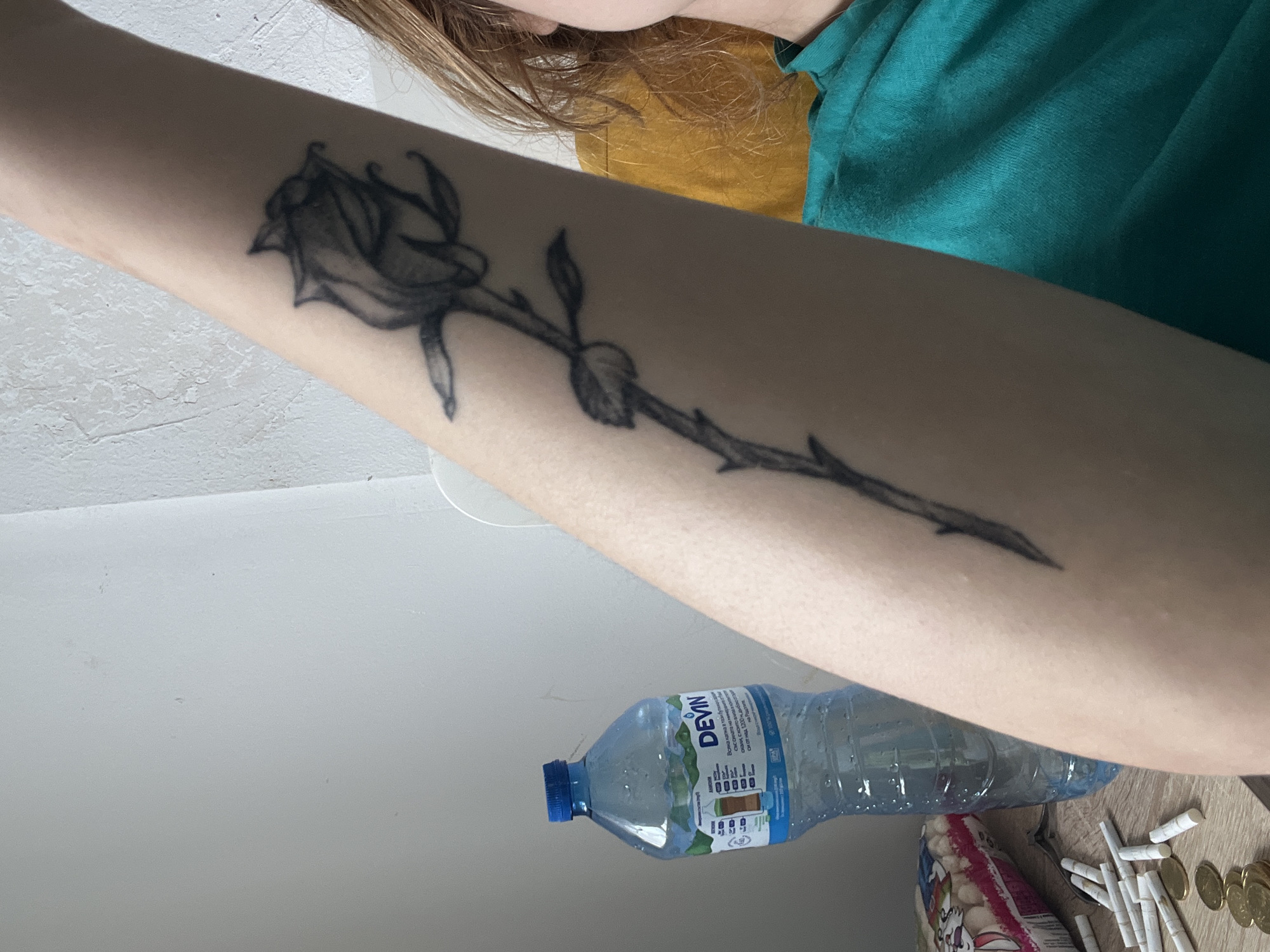A close-up shot reveals a person's left forearm, adorned with a black rose tattoo. The flower is detailed with shading and appears to be a line-art rendition. The stem of the rose extends down the forearm, incorporating leaves and thorns. The person is wearing a green shirt. In the lower portion of the photo, a bottle of Devin mineral water is visible, alongside coins and what appear to be hand-rolled cigarettes or tobacco products. The surface underneath is a light brown wood-grain. The location and city of Varna, Bulgaria cannot be determined from this image.