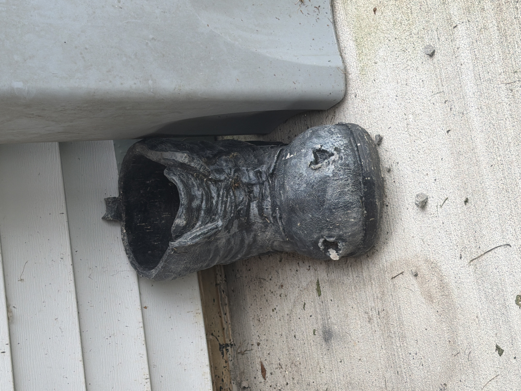 A black, weathered planter shaped like a boot sits on a concrete surface next to a light gray rectangular object. The boot planter is positioned vertically, with its opening facing upward and slightly to the left. It has a worn texture and shows signs of damage, including a hole in the toe area. The concrete surface is speckled with small stones and debris. Behind the planter and to the left is a section of white horizontal siding, possibly part of a building. To the right of the planter is the light gray object, which appears to be a large plastic container or bin. The scene is outdoors, with natural lighting. The image was taken in South Beaver Township, United States of America.