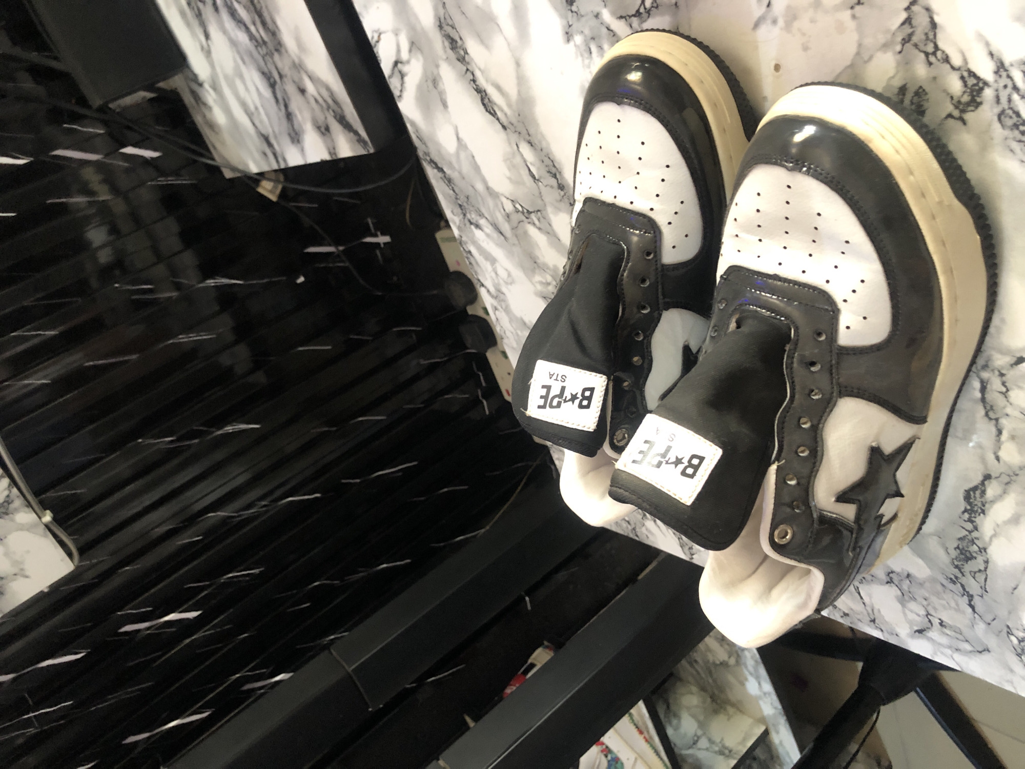 Two BAPE STA sneakers are displayed on a marble-patterned surface. The sneakers are a mix of black and white, with a black star logo on the side. The tongue of each shoe features a white label with the BAPE STA logo. The background is out of focus and appears to be a dark, slatted structure. The overall impression is of a product display.