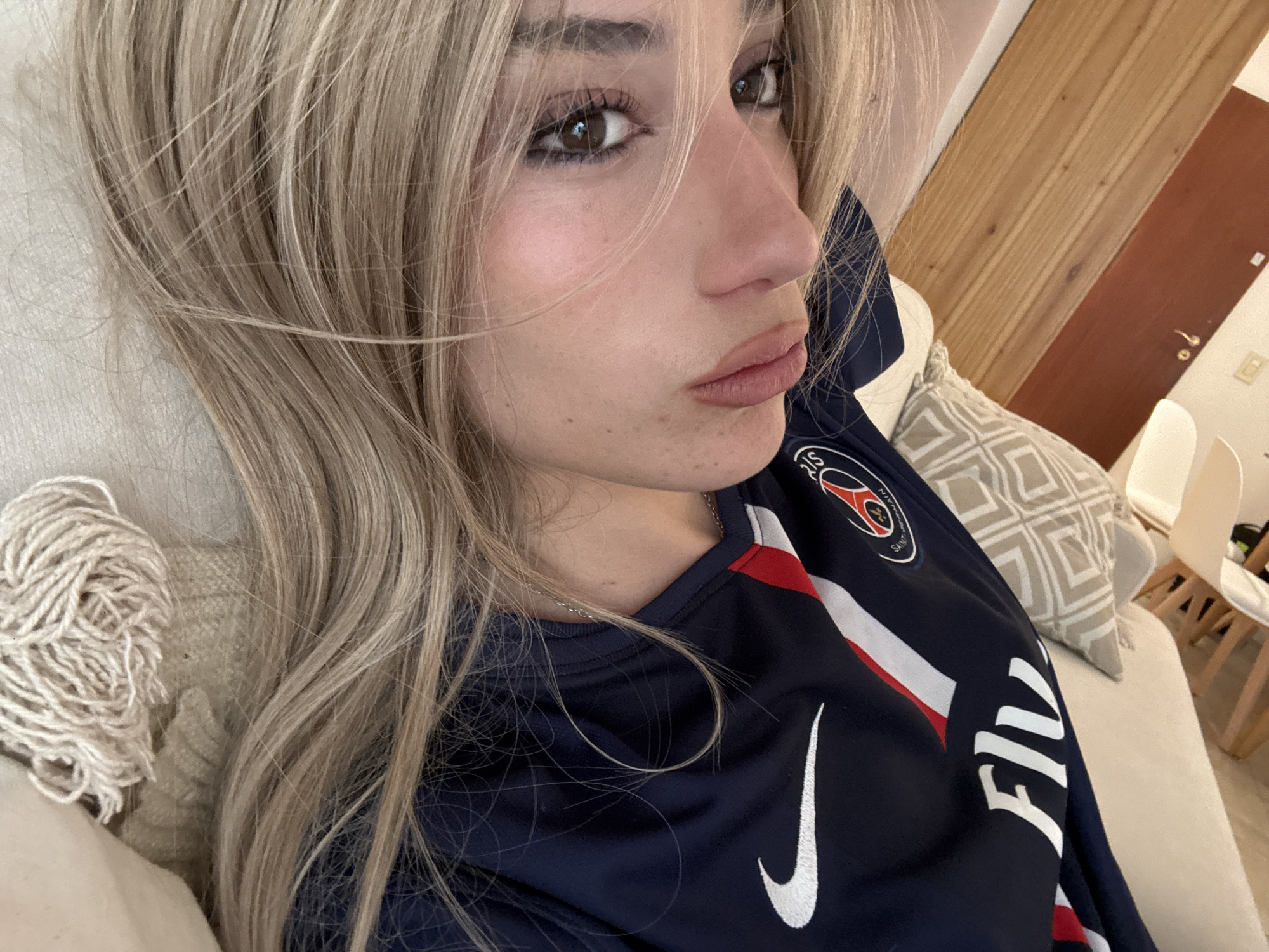A young woman with light blonde hair is lying down on a light-colored couch or bed, wearing a dark blue Paris Saint-Germain (PSG) soccer jersey with a white Nike logo and the word "FLY" in white lettering. She has brown eyes and is looking at the camera with a slight pout. Behind her are patterned cushions, a white chair, and a wooden door. The location cannot be definitively determined to be in Buenos Aires, Argentina from the given image.