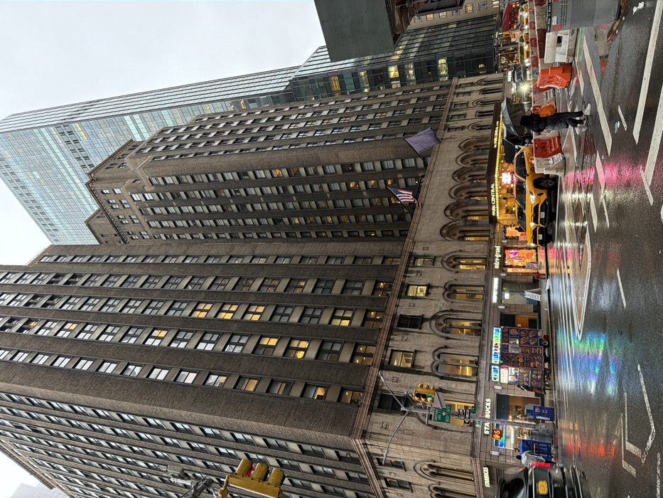 The scene depicts a street view at the intersection of West 55th Street and 7th Avenue in Midtown Manhattan, New York City, United States of America, during wet, likely rainy, conditions. The wet asphalt street reflects neon lights and building illuminations.
Dominating the frame are two tall, brown brick buildings with numerous rectangular windows, characteristic of older Manhattan architecture. The building on the right, identified by a prominent sign as "PARK CENTRAL," features an American flag and a purple flag displayed above its arched ground-level facade. Behind and to the right of these structures, a modern, towering glass skyscraper is visible under an overcast sky.
At street level, the road features white crosswalk markings and white traffic lane dividers. A traffic light assembly with active green and red signals hangs over the intersection. On the right side of the street, two yellow Toyota RAV4 taxi cabs are parked, facing away from the viewer. Orange and white traffic barriers are positioned in the street to the right of the taxis.
Several individuals are present: one person wearing dark clothing and white shoes holds a black umbrella while crossing the street from right to left. Another person stands near a food cart on the left sidewalk. The food cart, featuring a blue awning, displays various food items. Multiple storefronts are visible along the building's base, including "Starbucks" and "Miré-Grand," with their interior lights illuminated. A dark vehicle is partially visible on the far left of the frame.