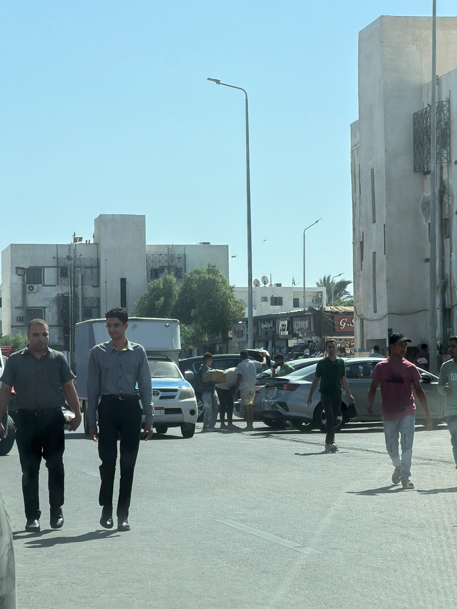 The image depicts a street scene in Sharm El Sheikh, Egypt, on a bright, sunny day. In the foreground, two men are walking towards the viewer. The man on the left is wearing a dark grey short-sleeved shirt and black pants. The man on the right is wearing a light blue long-sleeved shirt and black pants. Behind them, several cars are parked and driving on the street. Further back, a white building with multiple windows and balconies dominates the background, with another building to its left that appears to be an apartment complex. Several trees and streetlights are also visible. The sky is clear and blue.