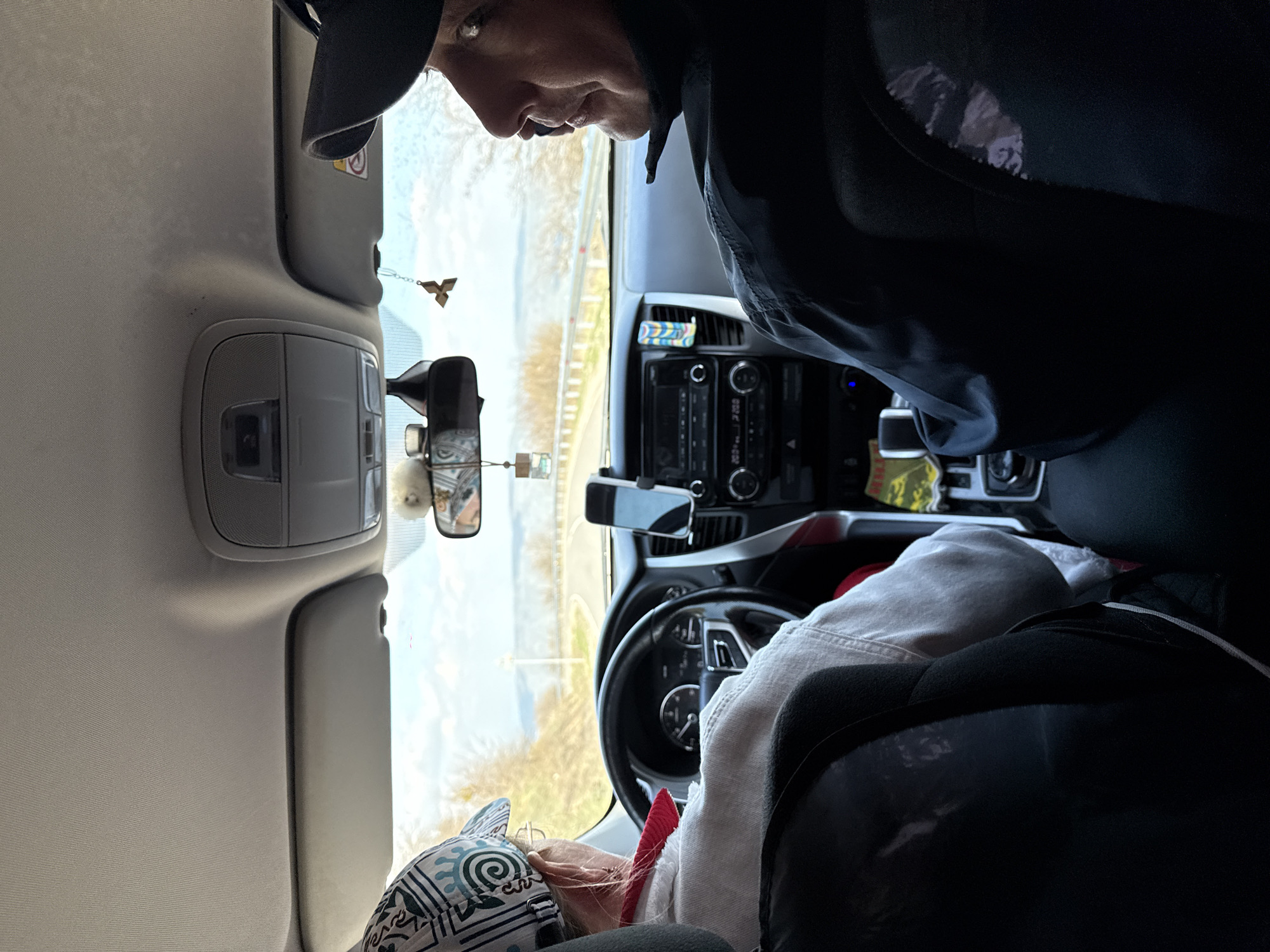 The image shows the interior of a car, possibly a van or SUV, with two people visible. One person, wearing a black jacket and cap, is leaning over towards the driver's seat. The other person, wearing a patterned hat and a light-colored jacket, appears to be lying down or reclining in the driver's seat. The car is on a road, and a landscape with trees and possibly a road are visible through the windshield. The rearview mirror has decorations hanging from it. The car's dashboard and controls are visible. The location of the image is assumed in the city of Novo-Zubutli, Russia.