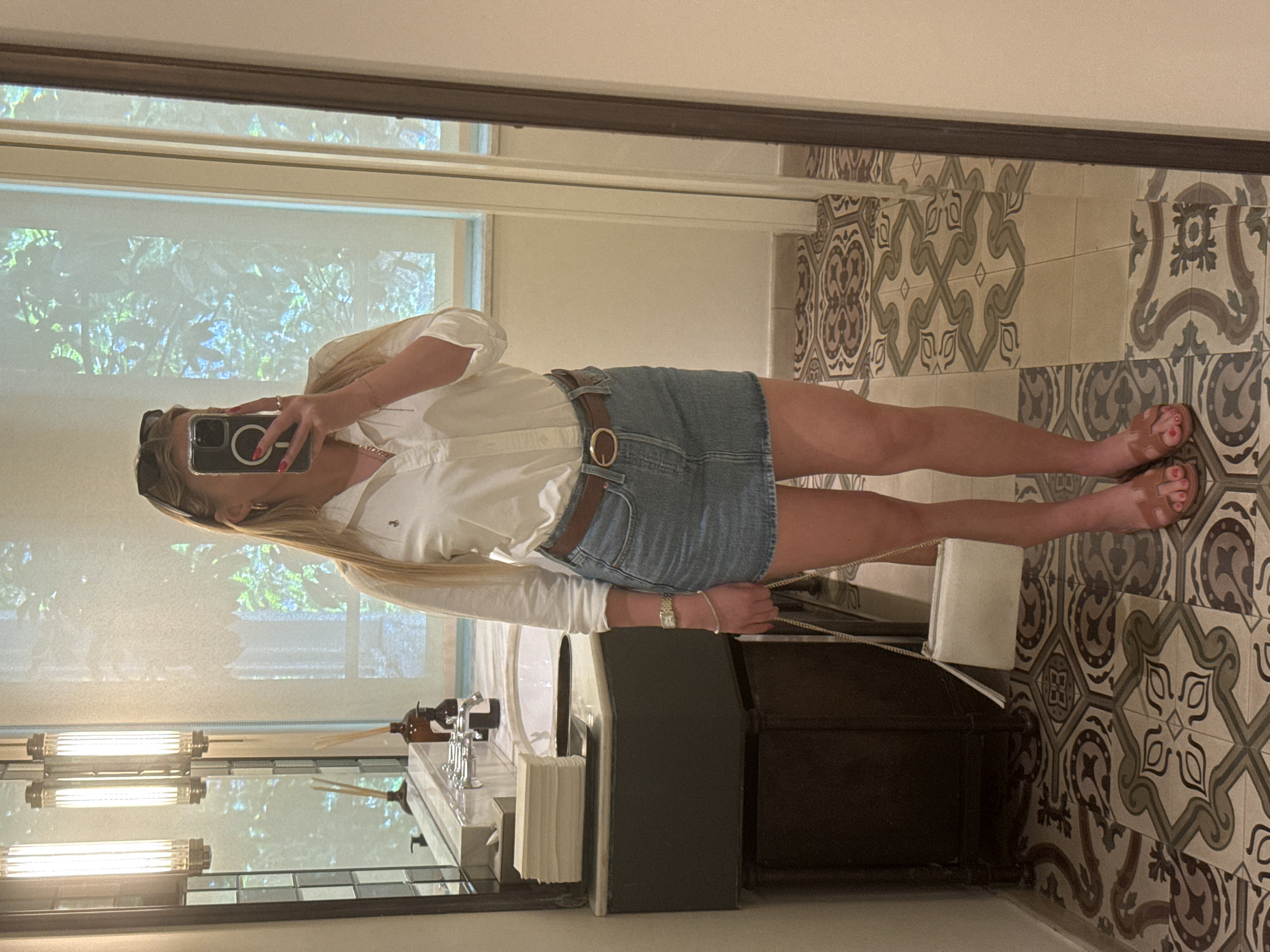A reflection in a mirror shows a woman holding a cell phone, presumably taking a selfie. She is wearing sunglasses on her head, a white, long-sleeved shirt, a denim skirt with a brown belt, and sandals. A bracelet and a watch are visible on her wrist. The reflection includes a bathroom scene with patterned floor tiles, a vanity with a sink, toiletries, and overhead lights. A window is behind her, displaying greenery. The city is Unknown, Unknown.