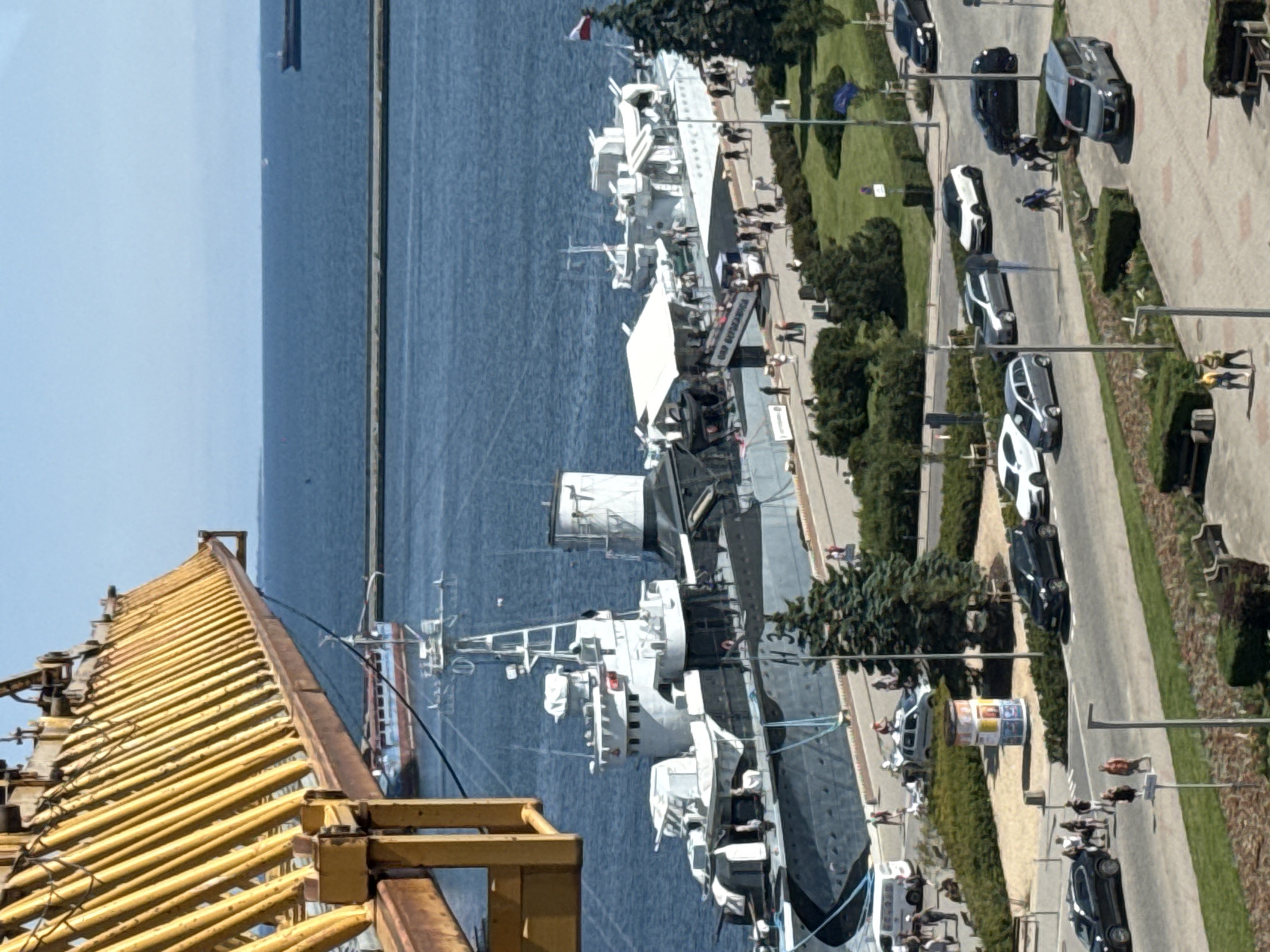 An aerial view shows the ORP Błyskawica museum ship docked in Gdynia, Poland, on a sunny day. The ship, a former Polish Navy destroyer, is gray and black with white upper structures and is adorned with a Polish flag at its stern. A large yellow crane, likely part of the port's infrastructure, dominates the left side of the frame, casting a shadow over part of the ship.
The ship is situated along a promenade where numerous people are visible walking around and disembarking from the ship via a ramp. Some are clustered near stalls or information booths. The promenade is lined with green trees and landscaping.
Beyond the promenade, a street is visible with several cars parked and driving, including a white van, black sedans, a gray hatchback, and a white SUV. Pedestrians are also present on the street and sidewalks.
The background features the calm, blue waters of the sea, with a breakwater or pier stretching across the middle distance. Further out, another ship can be seen in the distance. The sky is clear and blue, indicating fair weather. The overall scene depicts a bustling maritime location with historical significance, as indicated by the presence of the museum ship and port activities.