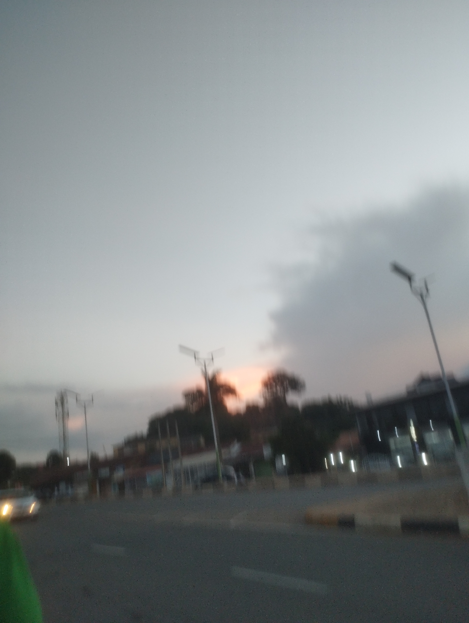 The image depicts an out-of-focus street scene at dusk in Jos, Nigeria. The sky is overcast with a gradient of grey, transitioning to a faint orange glow near the horizon, suggesting sunset or sunrise. Silhouetted against the sky are trees and buildings, some of which appear to be shops or commercial establishments with illuminated vertical lights. Several streetlights are visible, some with distinct T-shaped fixtures. A car with its headlights on is moving on the paved road, and a dark object, possibly a person wearing green clothing, is in the foreground on the left. The road appears to be a multi-lane thoroughfare with what looks like a median strip. The overall impression is of a somewhat busy urban environment at the end of the day, with limited visibility due to the blurriness and low light conditions.