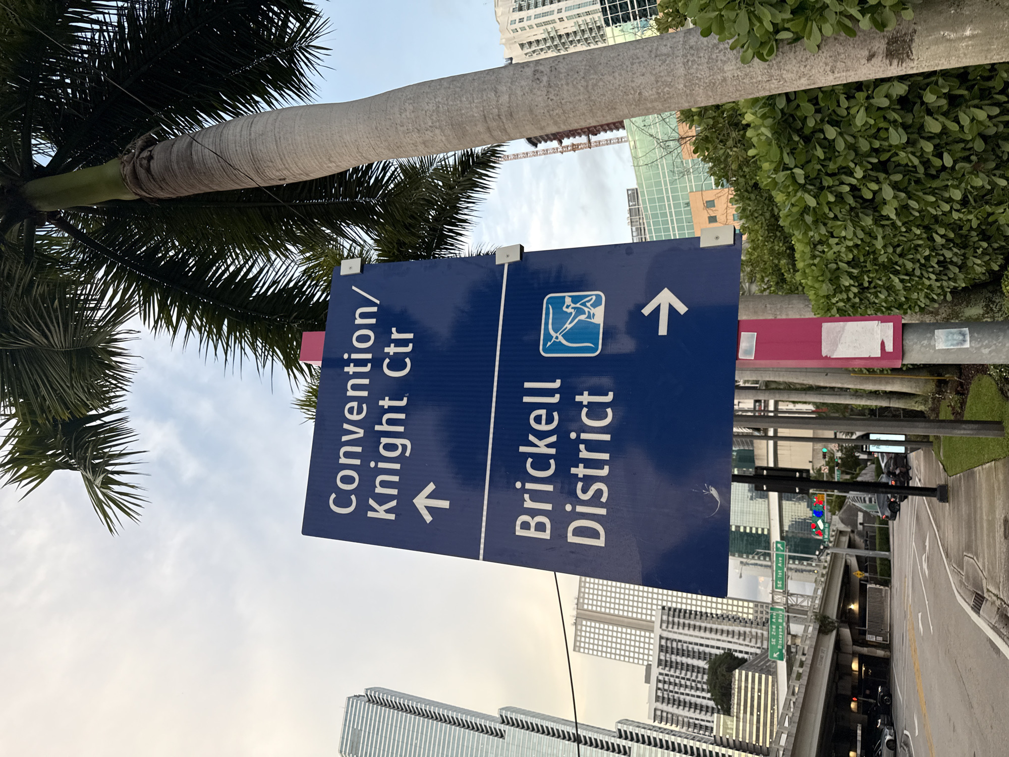 The image shows a street sign in Miami, Florida, USA. The sign is blue and directs people to the Convention/Knight Center and the Brickell District. The Convention/Knight Center is indicated with an arrow pointing to the left, while the Brickell District is indicated with a symbol and an arrow pointing to the right. The sign is located near a street with a sidewalk and a palm tree. In the background, there are buildings and a bridge.