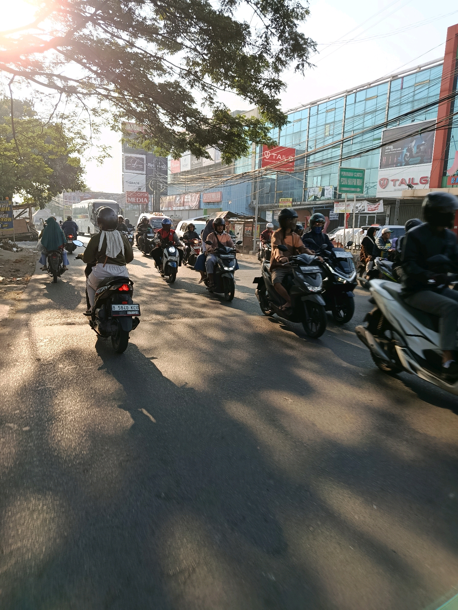 The image depicts a street scene in Bekasi, Indonesia, likely during the daytime. A traffic jam is evident, with numerous motorcycles forming a procession on a paved road. People on motorcycles are seen wearing helmets, with some wearing face coverings and casual clothing. The road is flanked by buildings, including commercial establishments with signage. A large tree with lush foliage partially obscures the upper portion of the frame, with sunlight filtering through its branches, creating dappled shadows on the road. Electrical wires are strung across the scene, typical of an urban environment. The atmosphere appears to be one of daily commuting or movement within the city.