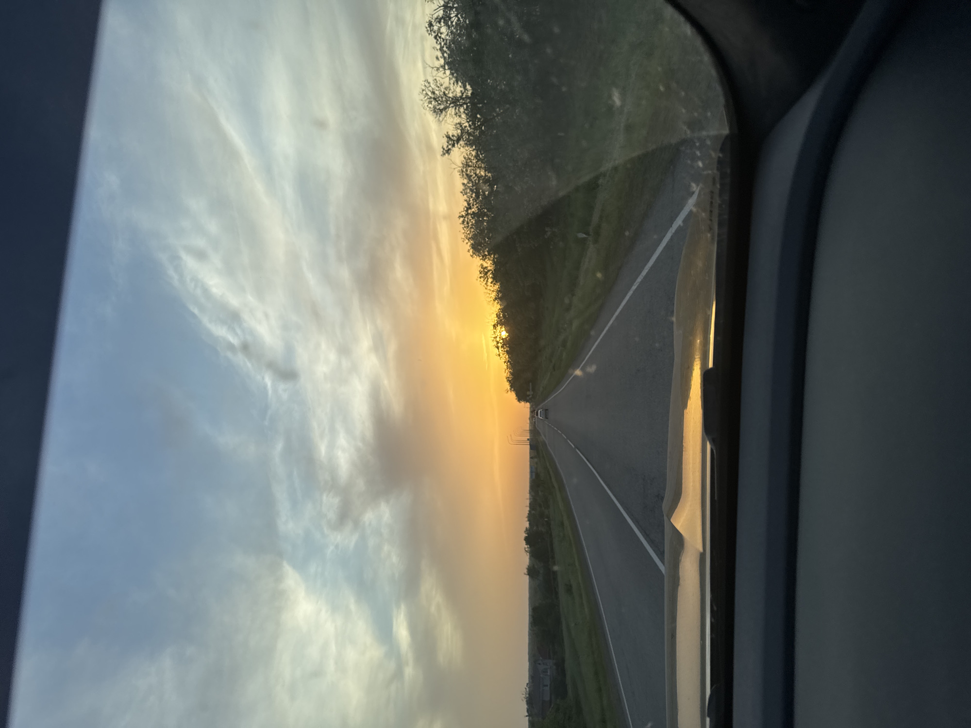 The photograph shows a view from inside a vehicle traveling on a road. The sun is setting ahead, casting an orange glow across the sky and reflecting on the windshield. The road is straight, with a white line marking the lanes. The roadside features trees and vegetation. The interior of the vehicle is visible on the sides of the frame, indicating it was taken from within the vehicle. The specific location in Novorozhdestvenskaya, Russia, is not evident from the image. The view is facing towards the direction of the sunset. The image does not contain evidence of people.