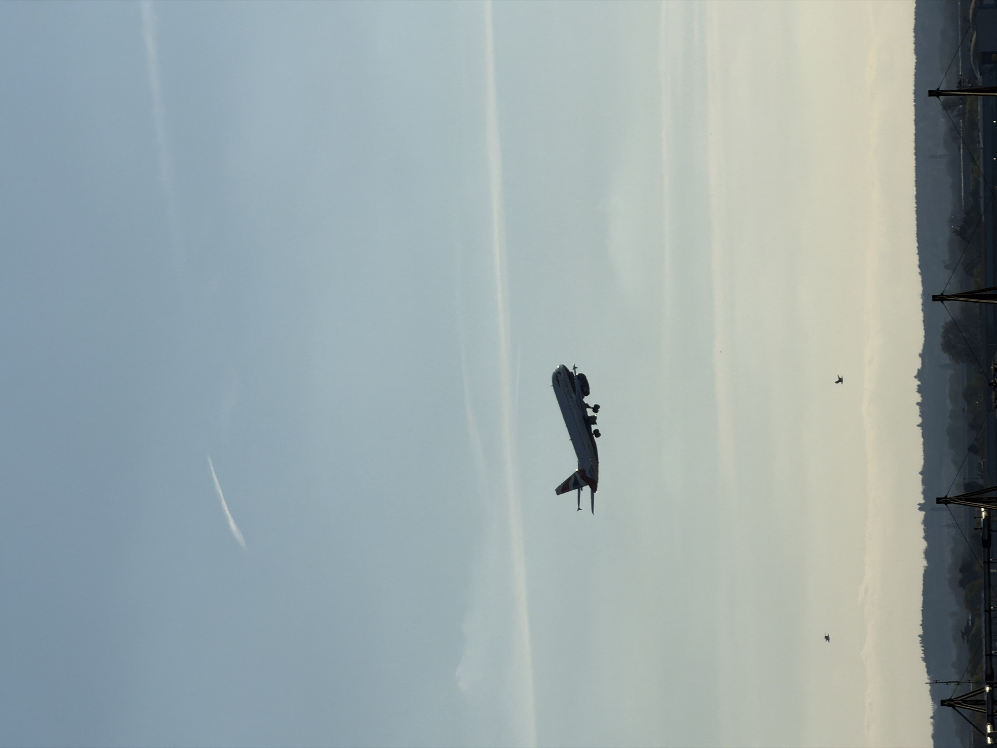 The image shows an airship flying in the sky. The sky is light blue with some clouds. In the lower right of the image is a treeline with some structures. There is one airplane or possibly two birds, to the right of the airship. The airship is white with red details. It is likely a sightseeing airship, and the location is likely London, United Kingdom.