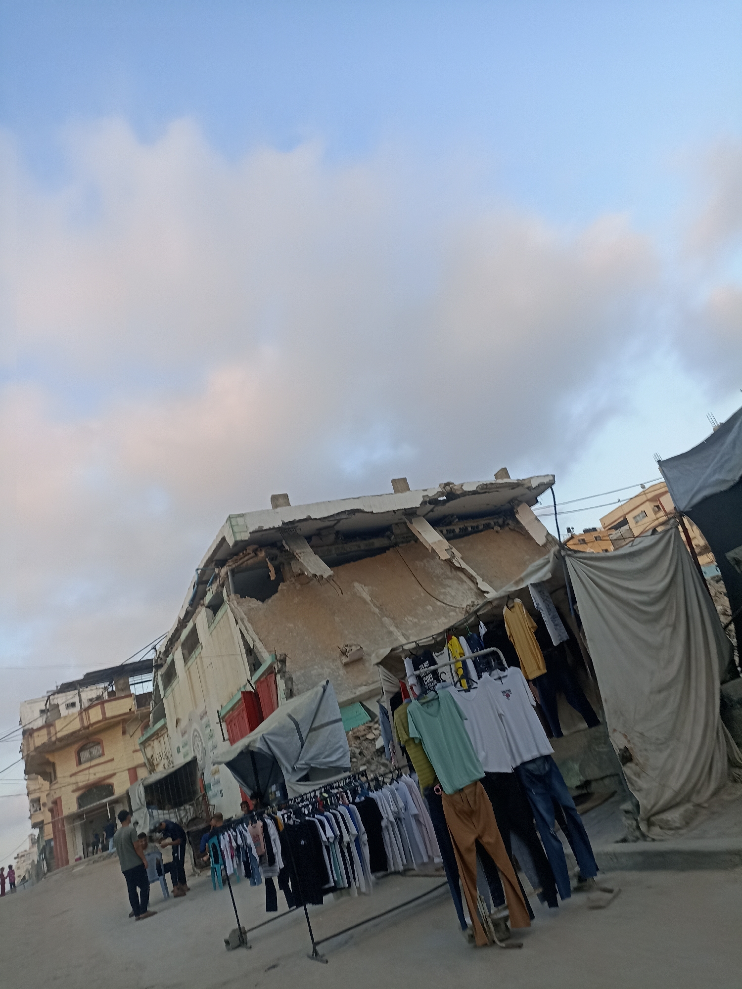 This media file depicts an outdoor scene in Gaza, Palestinian Territories, under a sky with scattered clouds, suggesting it is either early morning or late afternoon. In the foreground, a street market is set up with racks of clothing displayed for sale. Several people are visible, some browsing the merchandise, others attending to the stalls. Among the clothing items are t-shirts, pants, and what appear to be traditional garments.
The backdrop of the scene features a damaged building, showing signs of structural distress with a section of its facade collapsed. Adjacent to this building are other structures, some appearing intact and residential, while others also show wear and tear. A large, draped tarp is set up, possibly for shelter or to cover goods. The overall impression is one of resilience and daily life continuing amidst signs of conflict or destruction. The mood appears to be a mix of commerce and the somber reality of the environment. No visible text is clearly legible.