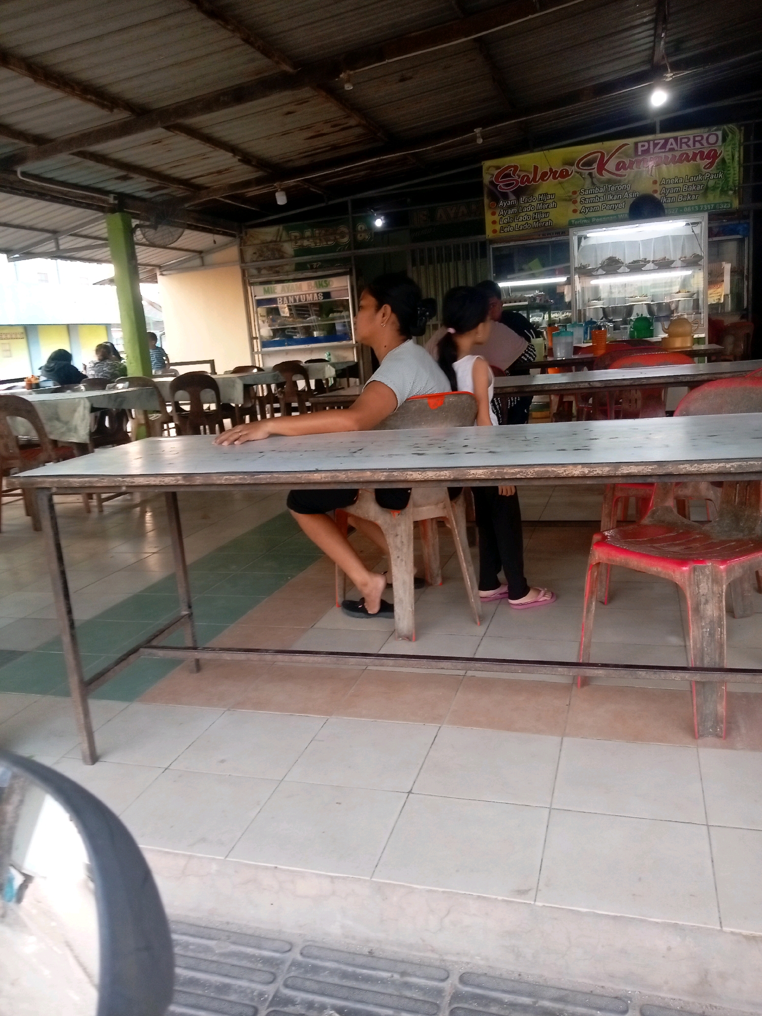 The image depicts an outdoor food court area, likely in Batam, Indonesia, given the signage visible. Two women are seated at a table, one in the foreground and another further back. The woman in the foreground is wearing a light-colored t-shirt and black pants, with her legs crossed. The other woman, partially obscured, appears to be a child wearing a white top and black pants. In the background, a banner displays "PIZARRO Salero Kampung" and lists food items. A display case filled with food is also visible, along with various tables and chairs. The environment appears to be a covered area with a corrugated metal roof supported by wooden beams and a green pillar. The floor is tiled. A car's side mirror is reflected in the bottom left corner of the image.