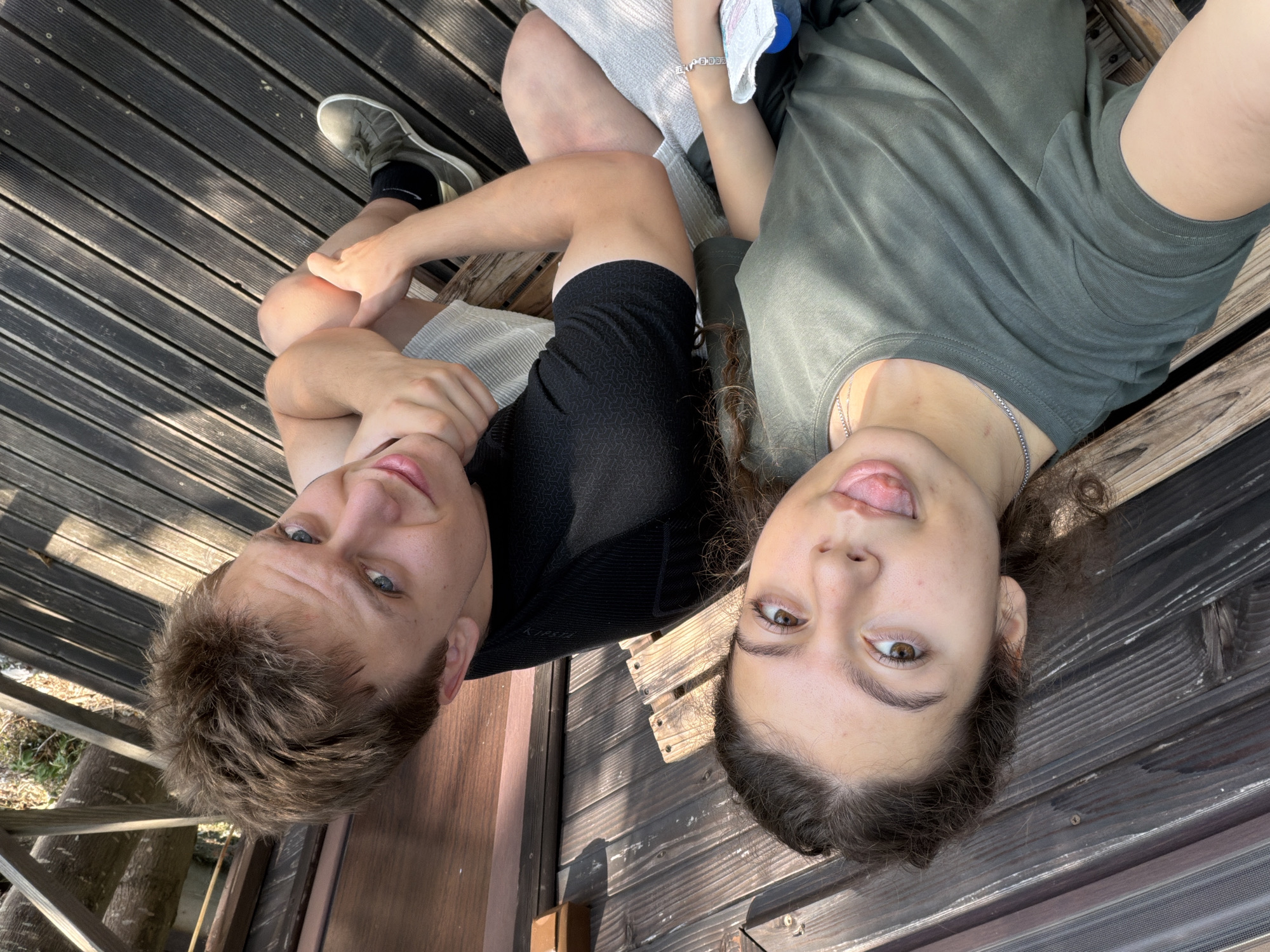 Two individuals, a young woman and a young man, are seated side-by-side on a wooden bench outdoors. The woman is in the foreground, positioned on the left, and is looking towards the camera with her tongue sticking out playfully. She has curly brown hair and is wearing a dark green t-shirt and a silver necklace. The man is to her right, looking towards the camera with a slight smile. He has short brown hair and is wearing a black t-shirt and shorts. They are seated on a wooden deck, with wooden walls and railings visible in the background. The scene appears to be a casual outdoor setting, possibly a park or a recreational area, with dappled sunlight filtering through the trees. There is no direct information about the location being in Apriltsi, Bulgaria.