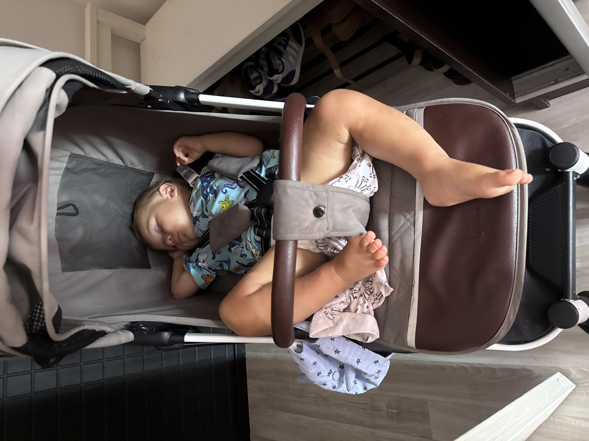 In a stroller located in Sochi, Russia, a young child is asleep. The child is wearing a blue t-shirt with a pattern of sea creatures. Their legs are extended, and a light-colored blanket with a star pattern is draped over their left side. A brown stroller bar is positioned in front of the child. To the right of the stroller, a shelf holds a pair of blue and white athletic shoes. The floor appears to be light-colored laminate. A black textured surface is visible to the left of the stroller.