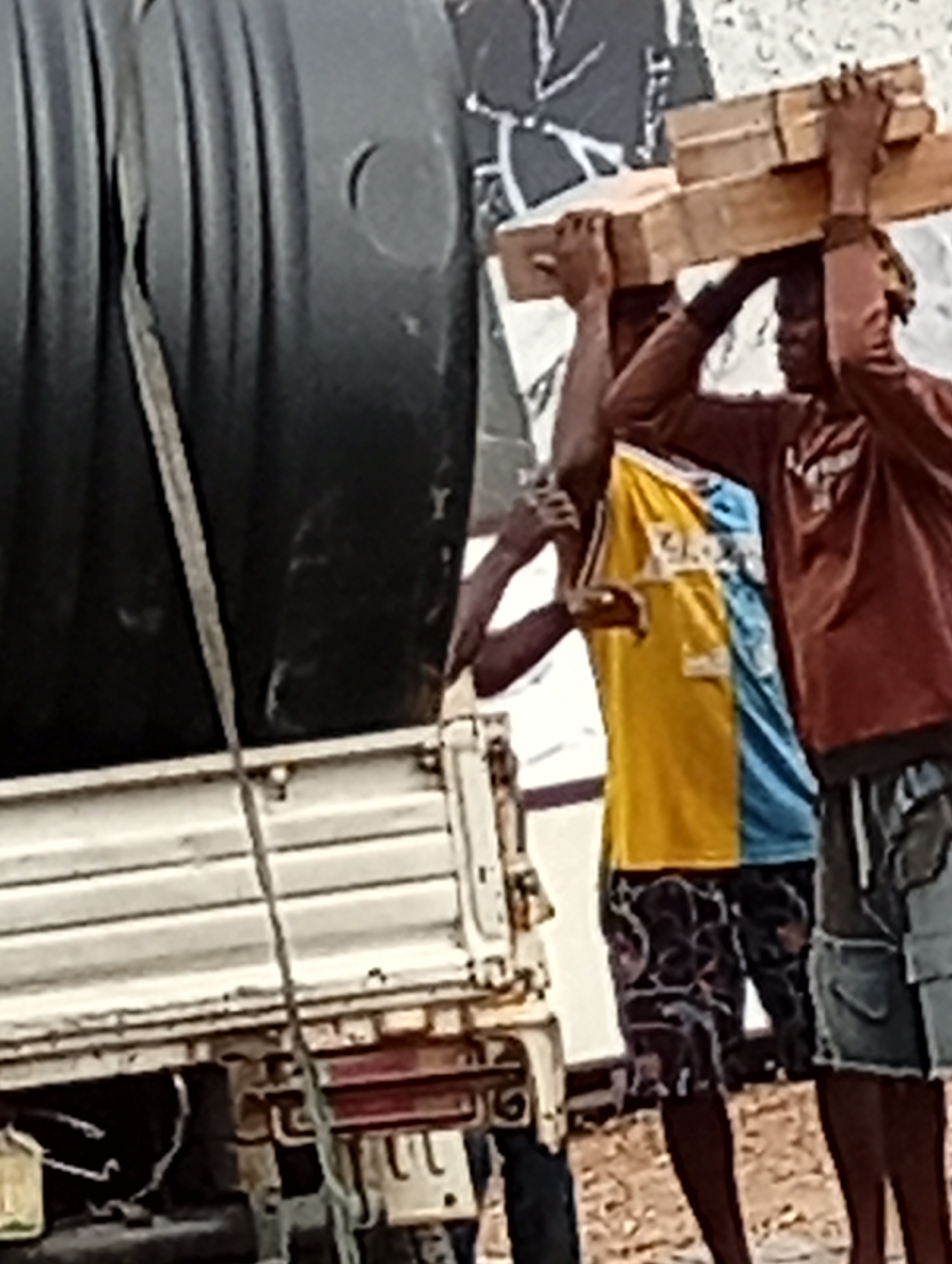 In Adankolo, Nigeria, a large black plastic water tank is strapped onto the back of a white flatbed truck. Three men stand next to the truck, one of whom carries a stack of wooden planks balanced on his head. Another man assists with the load, while the third stands nearby. The scene appears to be a delivery or unloading operation.