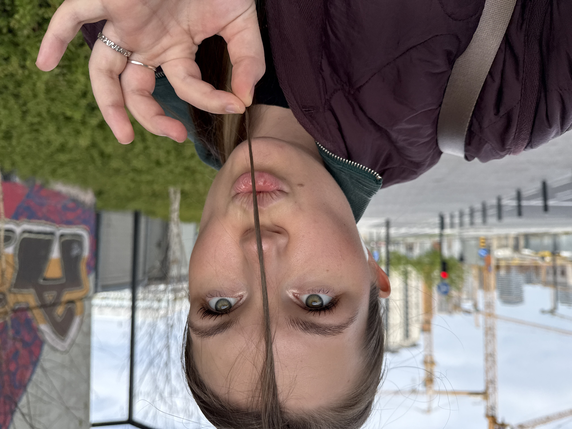 The image shows a woman holding a strand of hair between her lips and fingers. She is wearing a burgundy quilted jacket with a green collar and a shoulder bag strap. She has two rings on one of her fingers.
Behind the woman, there is a city landscape that features a building, a crane, a stoplight, and a reflective surface.
The city is Belgrade, Serbia.