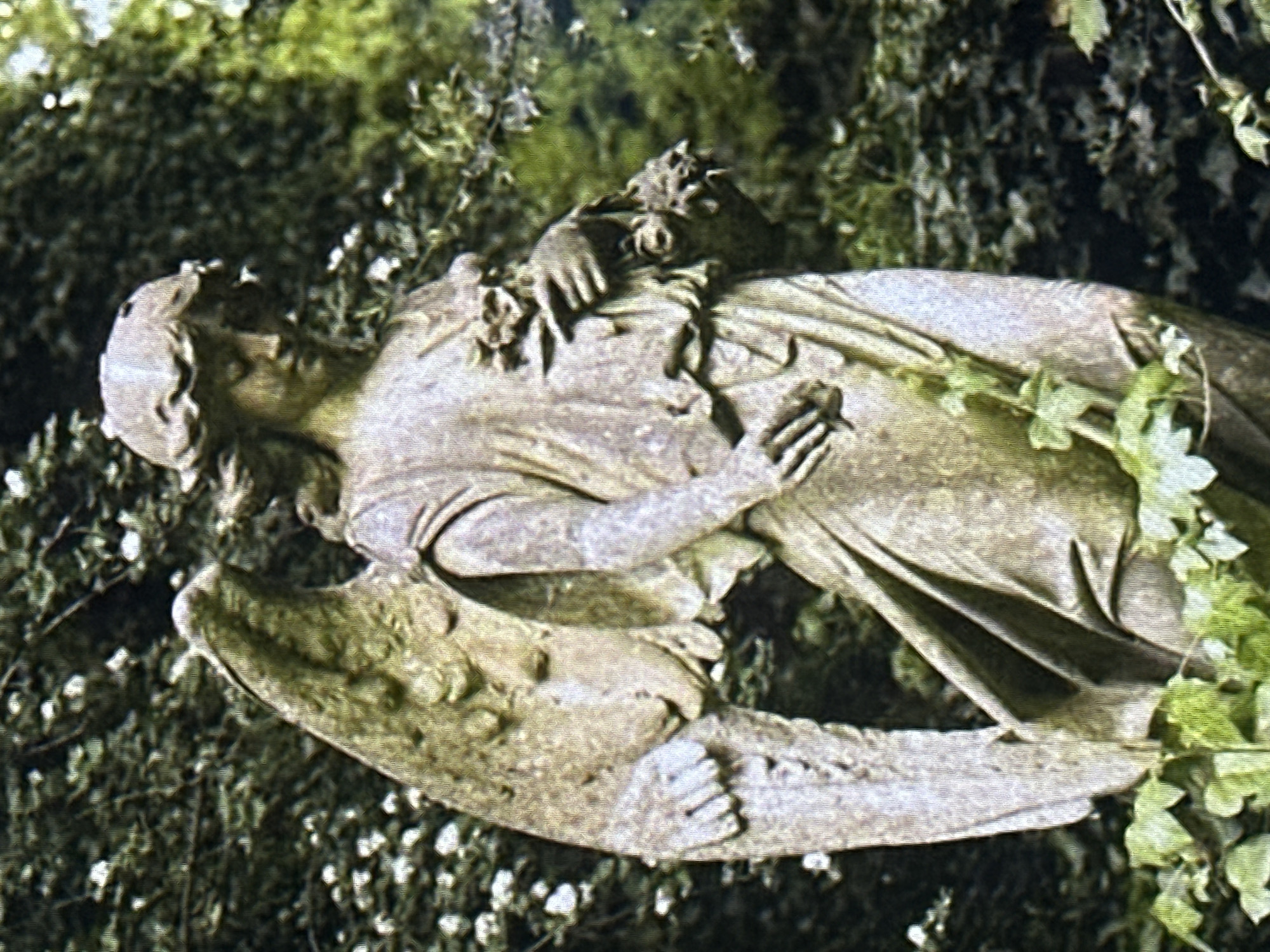 A weathered stone statue of an angel stands in a garden setting. The angel is depicted from the waist up, with one wing visible on the left side of the frame. It is wearing a draped robe and has a floral wreath on its head. The angel's right hand holds a bouquet of flowers, and its left hand rests near its waist. The statue is surrounded by lush green foliage, with some smaller white flowers interspersed in the background. The lighting suggests it is daytime, with soft shadows. The overall impression is one of a tranquil, possibly solemn, outdoor environment.