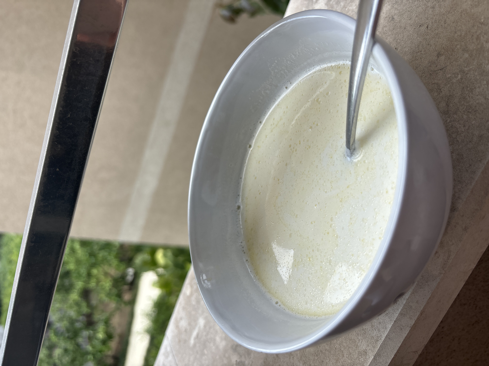 An overhead shot depicts a white bowl filled with a light-yellow, frothy liquid and a silver spoon. The bowl sits on a light brown, textured surface. To the left, a blurred metallic railing appears against a backdrop of green foliage. The location, based on the style of architecture and vegetation visible in the background, is likely an outdoor setting in Budva, Montenegro.