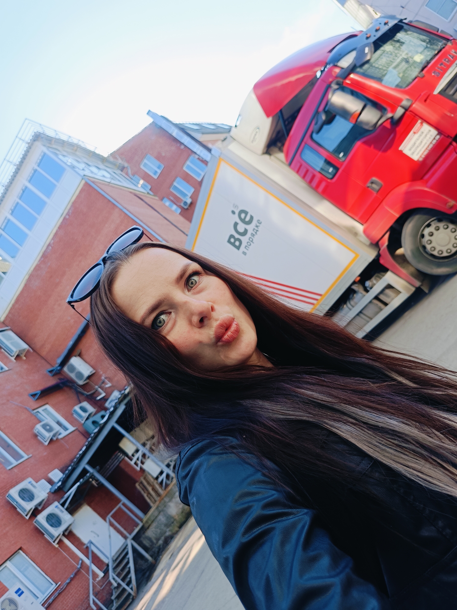 A tilted, eye-level selfie captures a young woman in the foreground, against an urban industrial backdrop in Chaikovsky, Russia. The woman has long, dark brown hair with lighter streaks, and dark sunglasses are perched on her head. She wears a dark, possibly leather, jacket. Her expression is playful, with lips pursed in a "duck face" and wide, blue-green eyes looking directly into the camera.
Behind her, a multi-story red brick building with numerous windows and many white air conditioning units is visible. To the right, a large red semi-truck dominates the background. The truck's cab is bright red, and the brand name "SITRAK" is discernible above the windshield. The attached white trailer features red stripes and prominent Russian text that reads "ВСЁ в порядке," translating to "Everything is in order." The truck appears to be parked near a loading dock or service entrance of the building.
The scene is brightly lit, indicating a clear, sunny day, likely in the late morning or afternoon. The sky is a light, clear blue. The overall setting suggests a commercial or industrial zone.