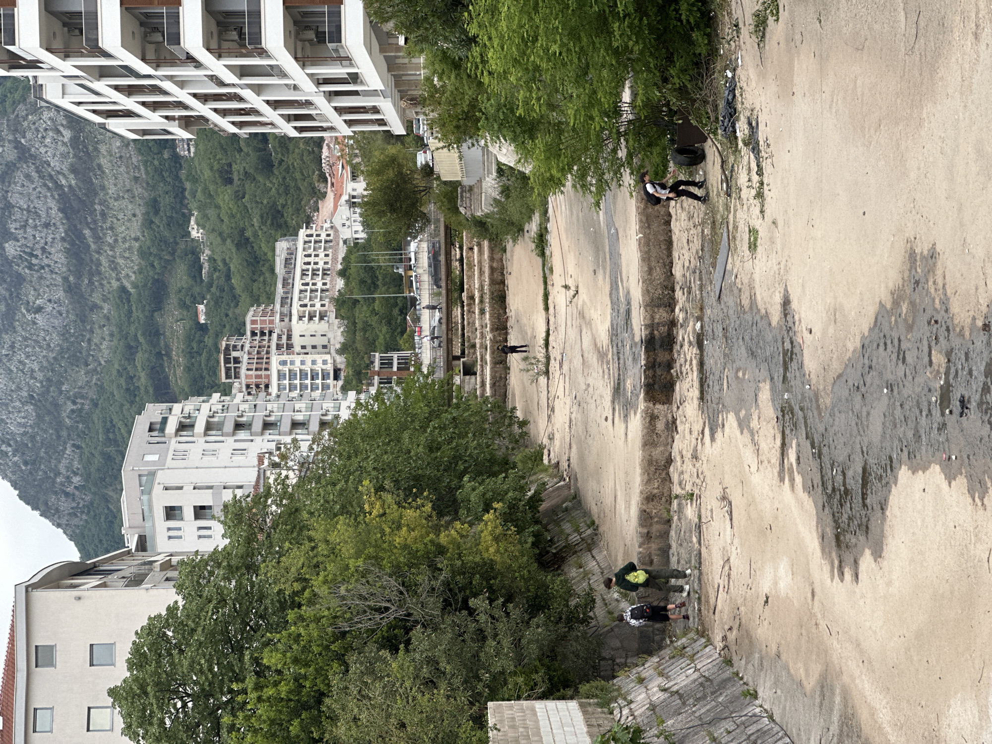 The image depicts a dilapidated concrete channel in Budva, Montenegro, with modern buildings and a tree-covered mountain in the background. Three people are visible within the scene, two near the bottom edge and one further down the channel. The foreground shows overgrown trees and bushes. The channel itself is stained with dirt and moisture, and appears neglected. A discarded tire sits on the edge of the channel near one of the people.