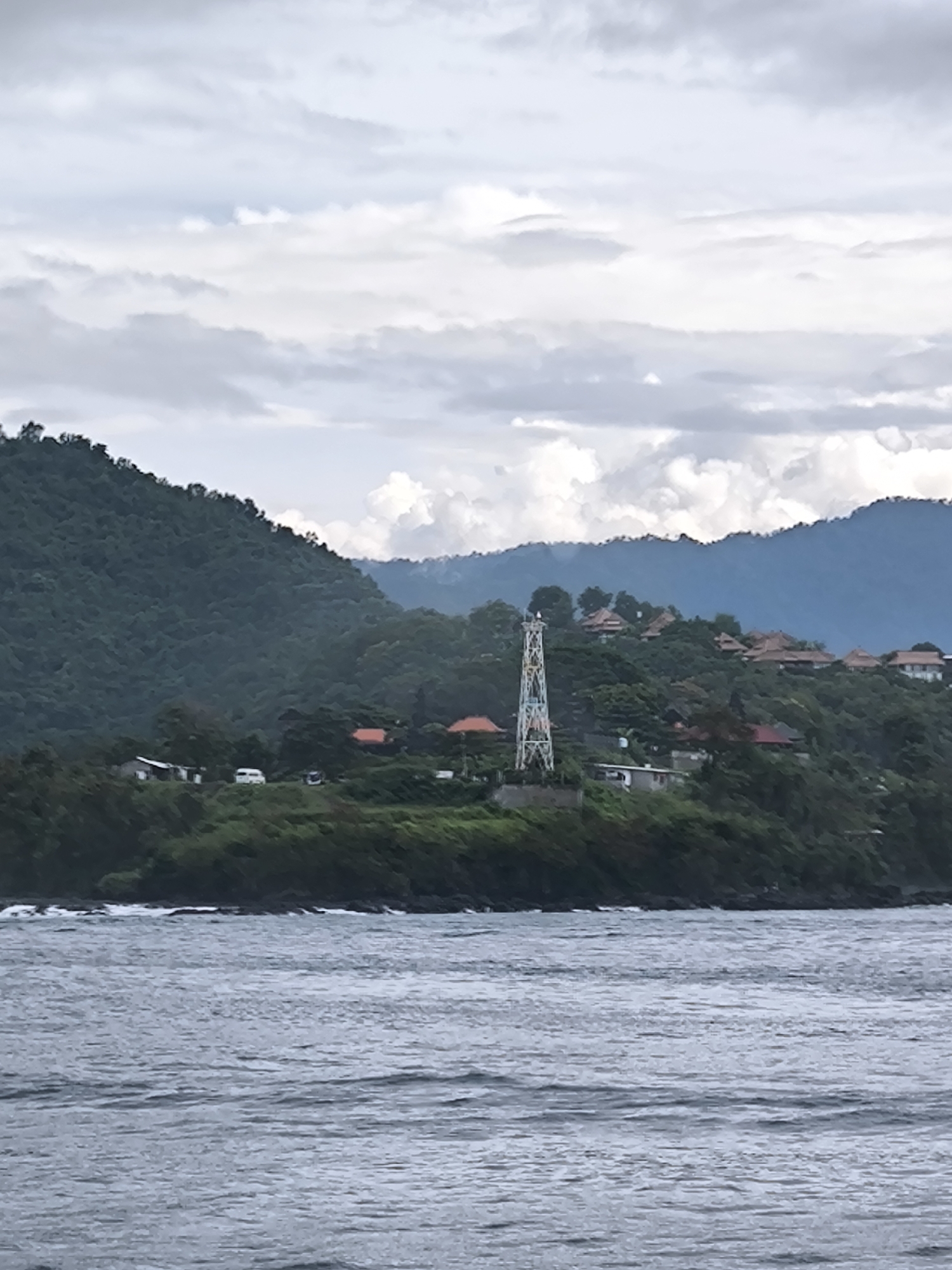 The image captures a tranquil coastal landscape viewed from the water, under an overcast sky. The foreground is dominated by choppy, dark grey-blue water, with small waves visible, particularly near the shore.
In the mid-ground, a lush green headland extends into the water, densely covered in tropical vegetation. A striking, tall, slender tower, possibly a lighthouse or communication mast, stands prominently on this headland. This tower is a notable feature, adorned with a vibrant, multi-colored mosaic-like pattern from base to top.
Scattered across the green hillsides, both on the headland and further up the slopes, are numerous buildings, predominantly featuring reddish-brown tiled roofs. These structures appear to be homes or resort units, nestled amongst the dense foliage. On the left side of the headland, some simpler buildings are visible, alongside what appear to be a white van and another vehicle on a winding road.
In the background, successive layers of large, hazy mountains, also covered in dense greenery, rise and fade into the distance, contributing to the sense of a vast natural environment. The sky above is filled with soft, diffused grey and white clouds, indicating an overcast day. The overall impression is one of natural beauty with subtle human integration, likely in a tropical or subtropical region. No specific human activity is in progress, and there is no visible text. The light suggests it is daytime, possibly morning or late afternoon.