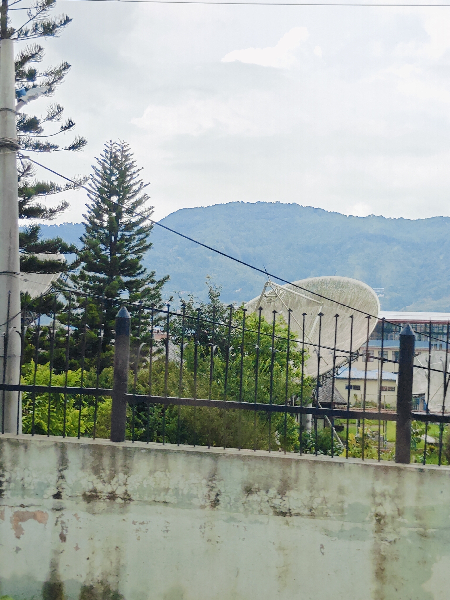 This is an outdoor, medium-shot, eye-level photograph taken in Chandragiri, Nepal. The image features a large satellite dish positioned behind a black metal fence, with lush green trees and foliage in the foreground. In the background, rolling hills covered in vegetation are visible under a cloudy sky. The satellite dish appears weathered, with a mesh-like surface. The fence is made of vertical bars with decorative tops, and it is supported by dark posts. Beyond the fence, several buildings with light-colored walls and dark roofs can be seen, along with more trees. The sky is overcast with patches of white clouds. A light pole stands on the left side of the frame, with wires extending across the scene. The foreground includes a concrete wall with a textured and stained surface. There are no people or animals present in the image.