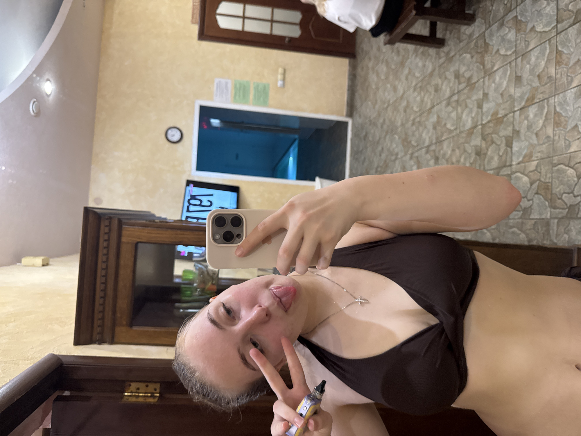 A female is taking a selfie in what appears to be a sauna. She is wearing a dark bikini top and a silver necklace with a cross pendant. She is holding an electronic cigarette in her left hand and making a peace sign. Her tongue is sticking out slightly. The sauna has wood-paneled walls and tile floors. There is a window showing a pool. A wooden cabinet is behind her. The clock shows 12:27. The location is in Chaikovsky, Russia.