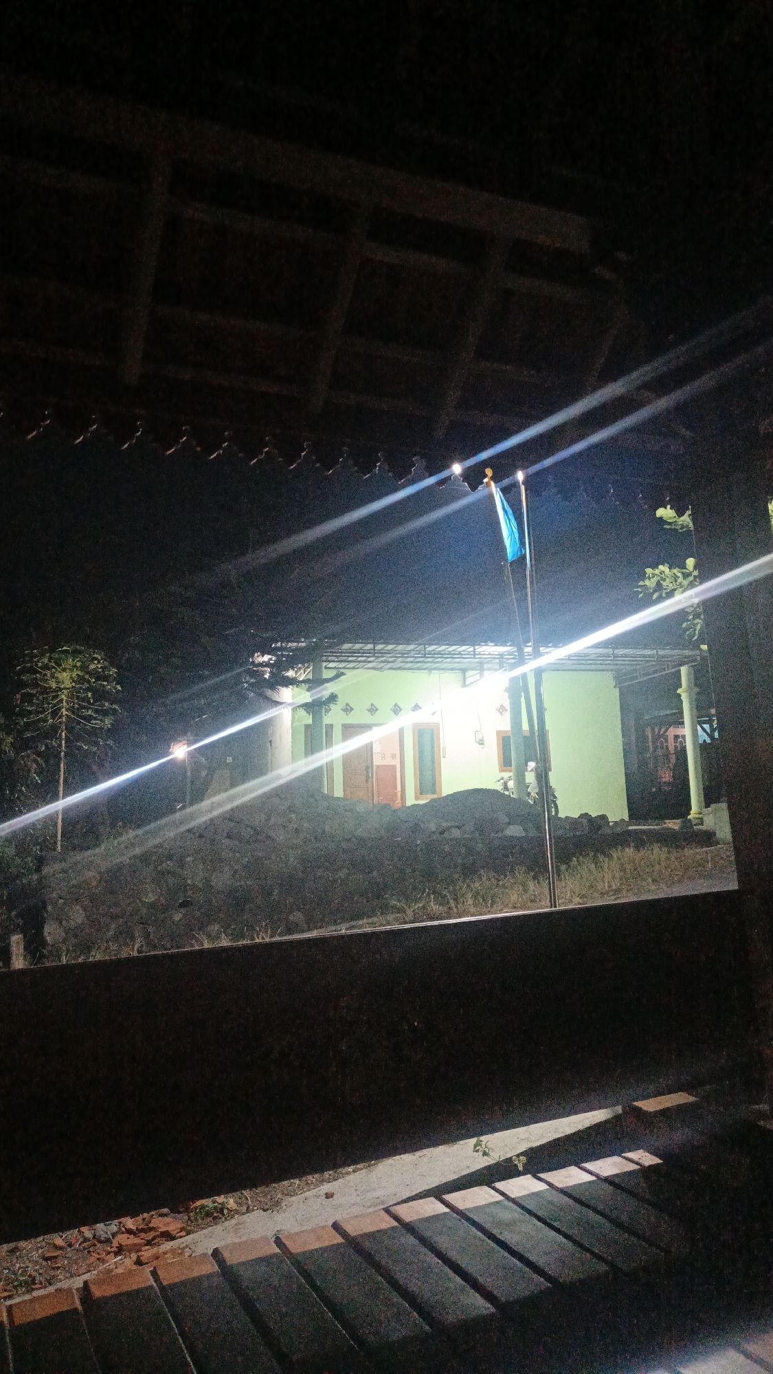 This is a nighttime photograph taken in the city of Sambi, Indonesia. The scene depicts a light green house with two doors and several windows, illuminated by outdoor lighting. A flagpole with a blue flag stands in front of the house. The foreground of the image is dominated by a dark, horizontal structure, possibly a fence or wall, and beneath it, a row of dark, rectangular objects, likely tiles or paving stones, are visible. The background consists of trees and a dark, uneven terrain. Intense light beams cut across the image, suggesting the presence of strong light sources and potentially causing lens flare. The overall atmosphere is dark and somewhat obscured by the lighting conditions and the overhanging roof structure in the upper part of the frame.