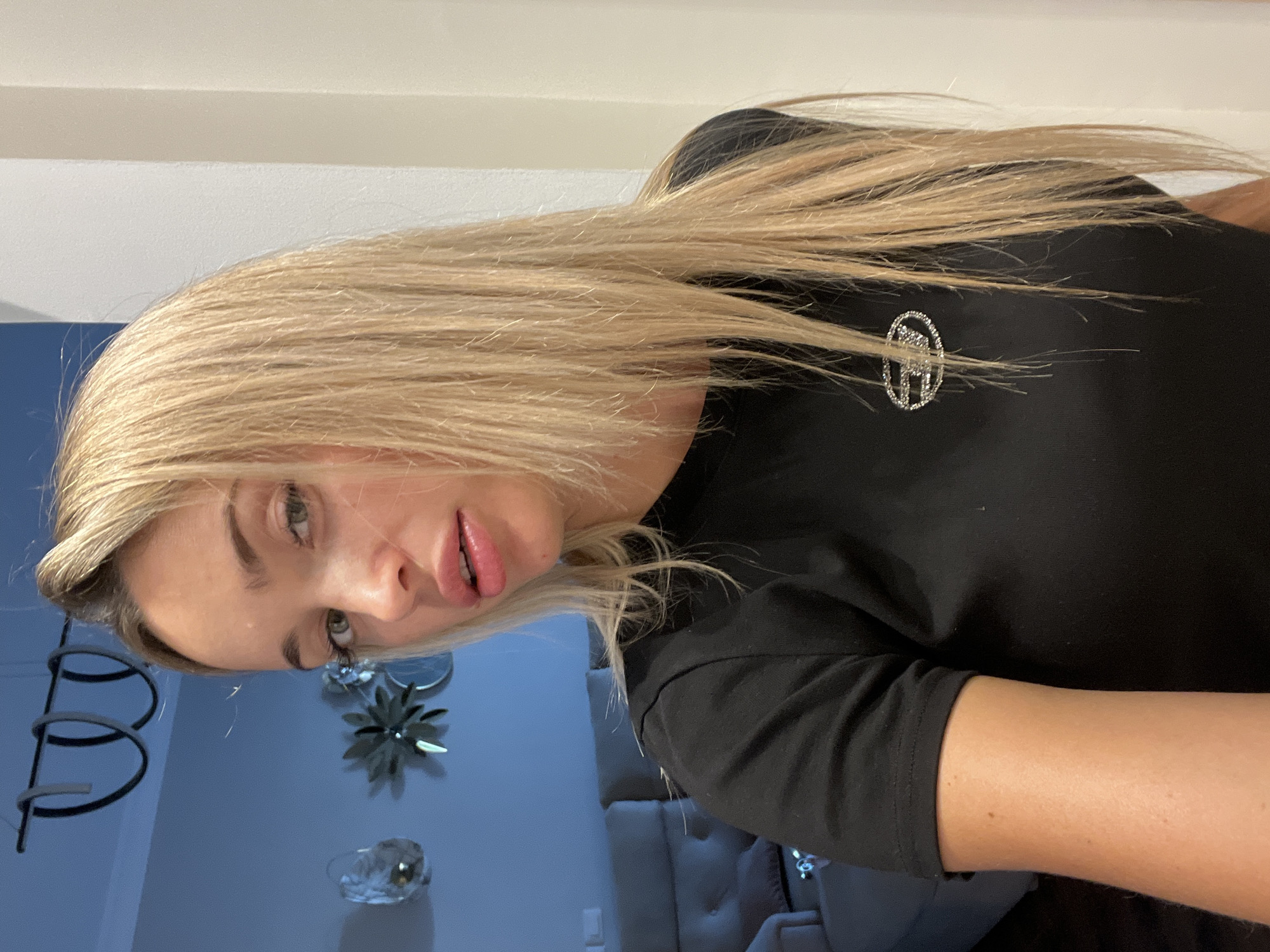 A young, fair-skinned woman with long blonde hair and green eyes is pictured indoors. She is wearing a black t-shirt with a small, oval-shaped emblem embellished with rhinestones on the left chest area. Her lips are slightly parted, and she is looking directly at the camera. The background shows a dimly lit room with a grey sofa, decorative wall hangings, and a white wall. The image is taken from a medium close-up angle, focusing on the woman's upper body and face.