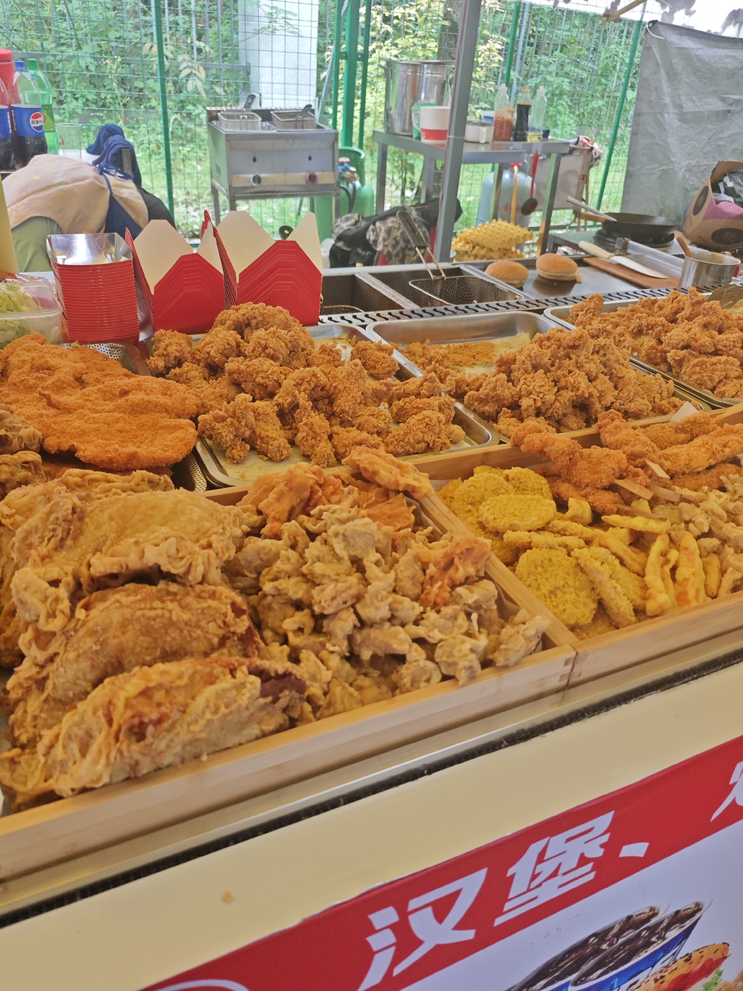 The image shows a food stall in what appears to be Shapingba District, China. The stall is selling a variety of fried foods. There are fried cutlets, fried chicken pieces, and various fried vegetables, including sweet potatoes and other root vegetables. Red boxes are stacked on the counter, presumably for take-away orders. In the background, there is a fryer and a grill, along with bottles of drinks and seasonings. The stall has a red banner with Chinese characters on the front, which seems to advertise the stall's offerings, including burgers (implied by the characters "汉堡"). The stall is set up outdoors, possibly in a park or public space, with a green fence in the background.
