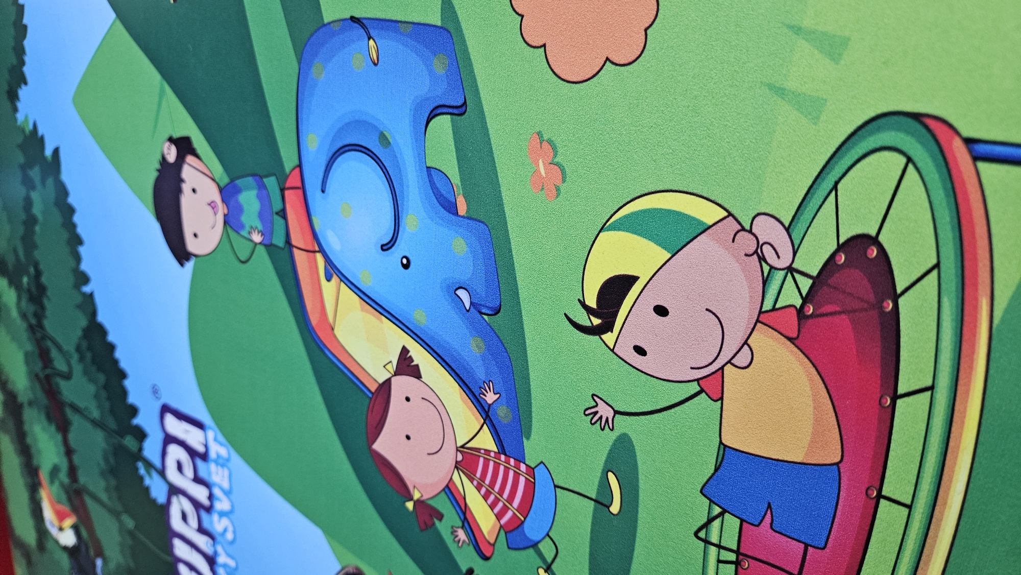 The image depicts a vibrant, cartoonish illustration with children and an elephant on a grassy hillside under a blue sky. In the foreground, a child wearing a yellow and green cap lies on a red and yellow amusement ride, waving. Behind this, a girl with pigtails in a striped shirt runs towards an elephant-shaped slide. Further back, a boy in a blue and green striped shirt stands on top of the slide. The background features trees and a distant landscape. The overall style suggests it is likely from a children's book, poster, or educational material. The text "UPPI" and "Y SVET" are visible in the upper left corner, suggesting a brand or title. This illustration is a graphic, not a photograph, therefore its location in Bratislava, Slovakia, cannot be determined.