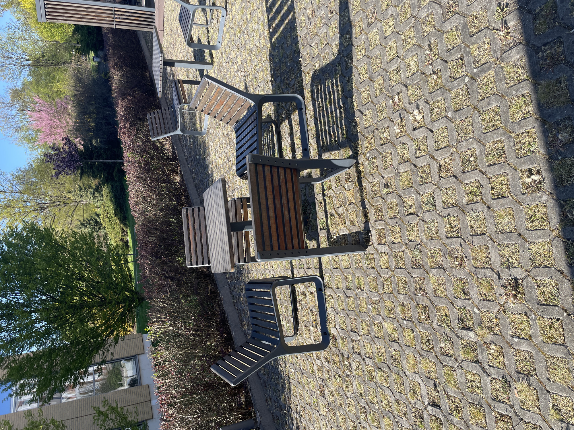 The image shows an outdoor setting with several picnic tables and chairs on a patterned brick surface. The furniture is modern, with wooden slats and metal frames. Adjacent to the paved area is a hedge, and beyond the hedge are trees, including one with pink blossoms. A building is visible in the background. The image is taken during daylight, with shadows cast by the furniture. The location appears to be in a park or a similar public outdoor space. There is no evidence to suggest this is in the city of Unknown, Unknown.