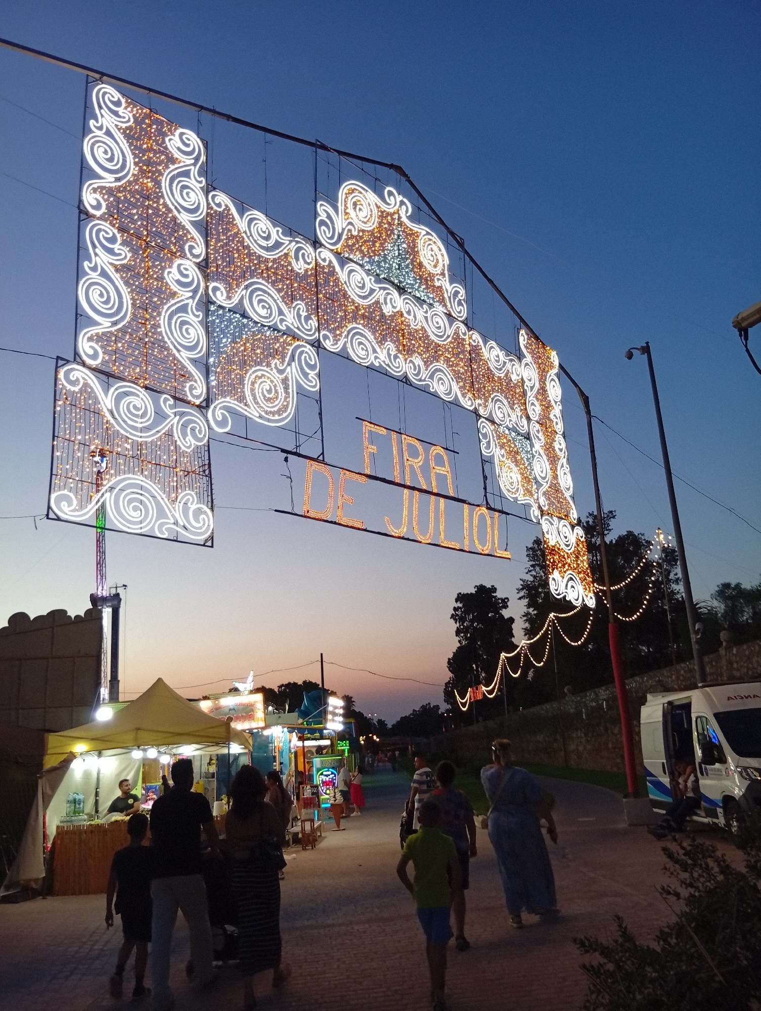 The image depicts a street scene at dusk in Valencia, Spain, likely during the "Fira de Juliol" festival. An overhead archway adorned with illuminated white and orange lights spans across the street, displaying the text "FIRA DE JULIOL" in orange neon letters. Various stalls are set up along the right side of the pathway, with people milling about. One stall has a yellow awning and appears to be a food vendor, with a person behind the counter. Further down, other stalls are visible, some with games and merchandise. A white van, possibly an ambulance, is parked on the right side of the road. A group of people, including families with children, are walking along the paved pathway. Trees and vegetation are visible in the background, silhouetted against the twilight sky. The overall atmosphere suggests a festive evening event.